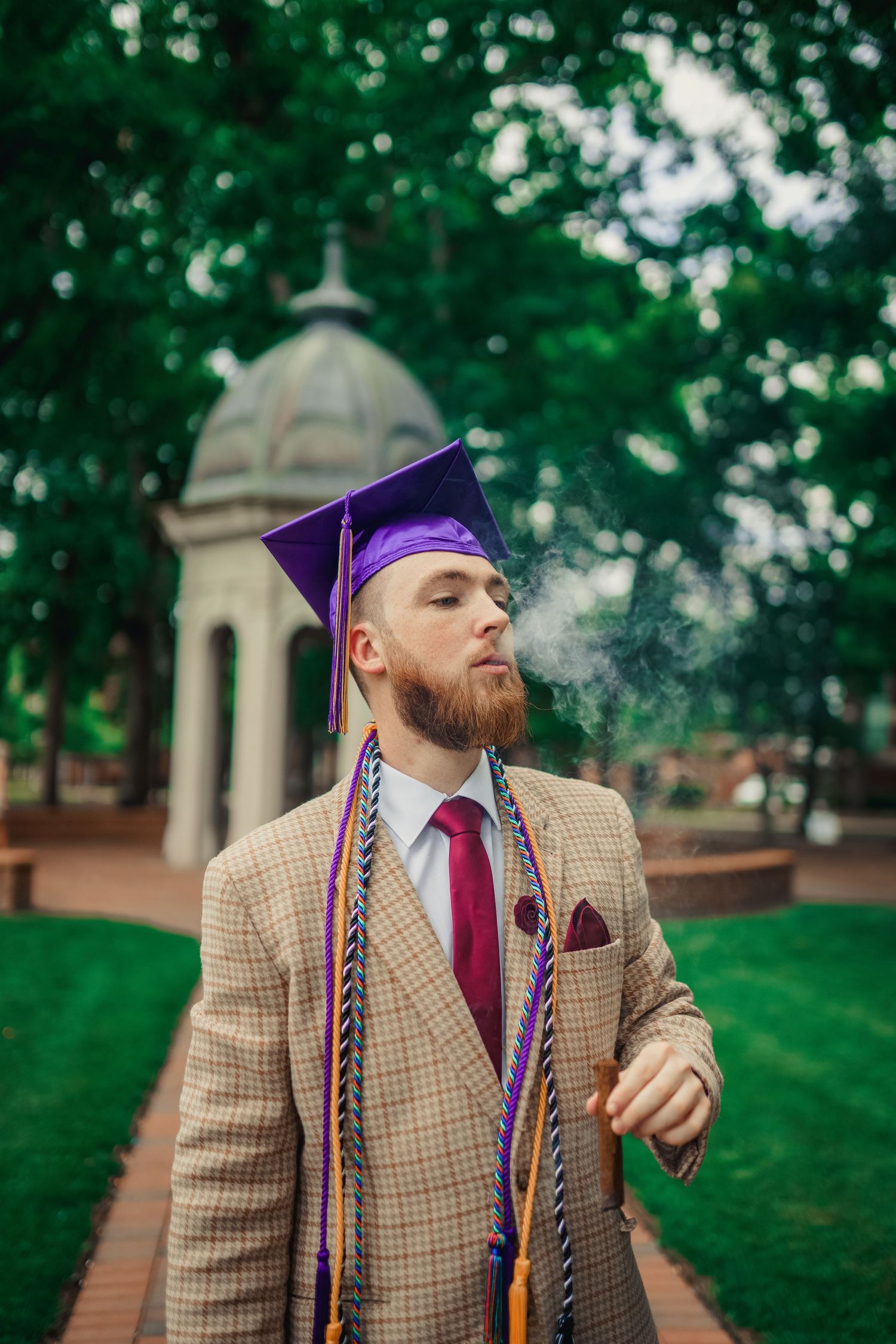 Graduate in purple cap and tan plaid blazer poses exhales cigar smoke while at the Cupola during his graduation photos