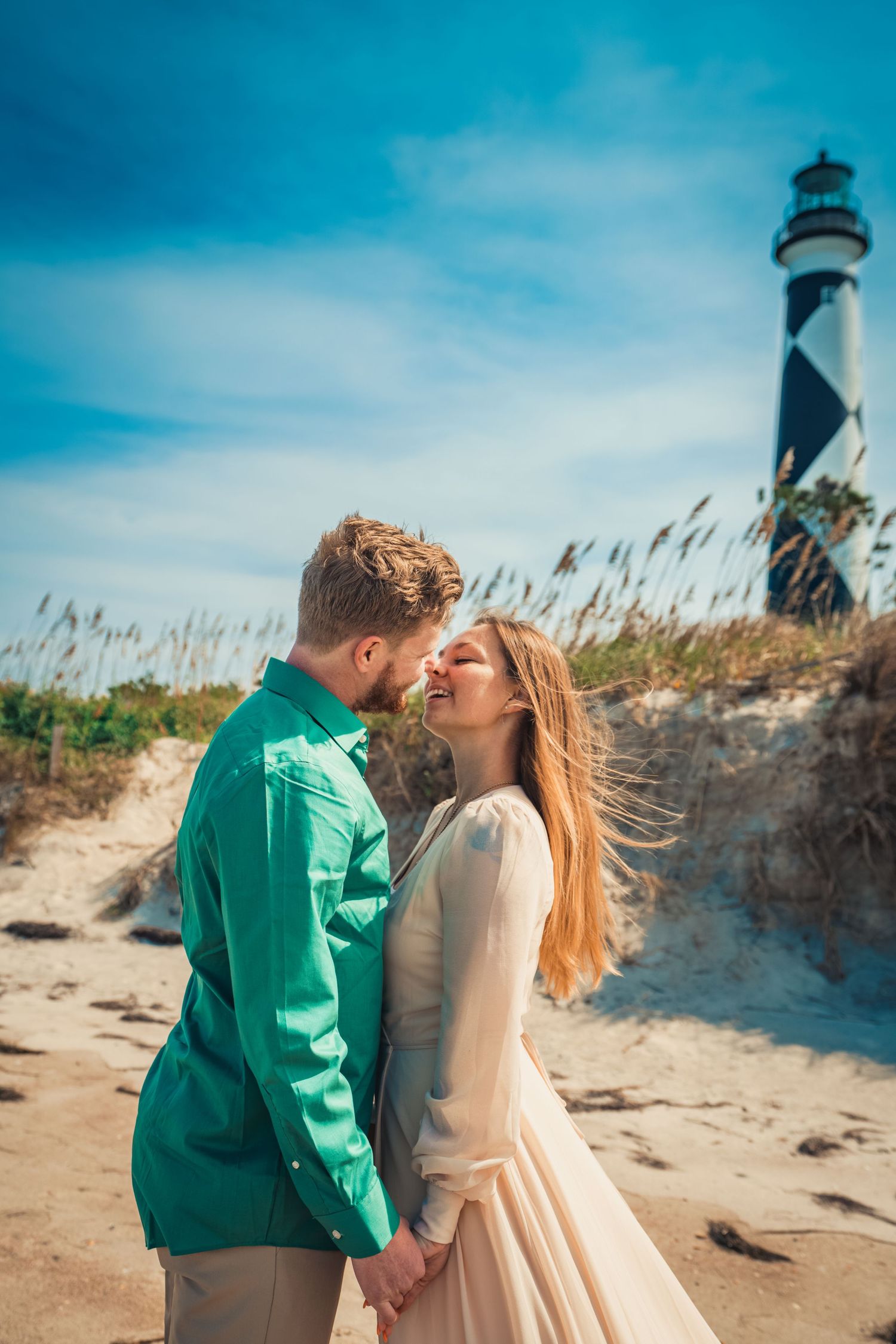 A couple shares an intimate moment on a sandy beach with a lighthouse and sea grass in the background.