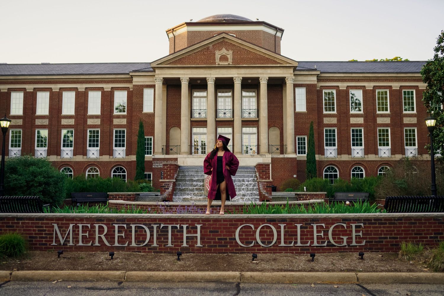 Graduate in maroon regalia poses in front of Meredith College main building entrance.