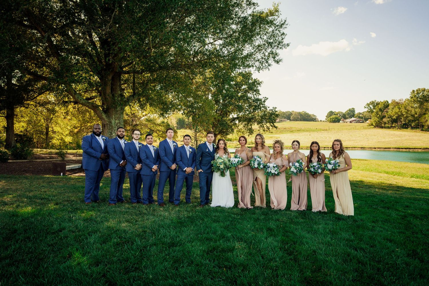 Wedding party in navy blue suits and beige dresses poses for formal photo on lush green lawn at sunset.
