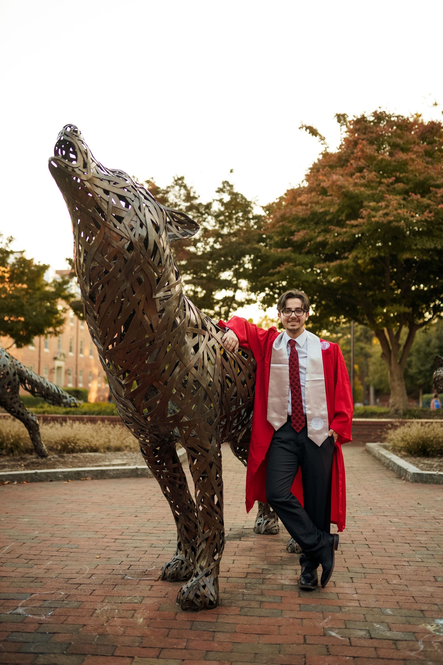 Graduate poses with metallic wolf statue on brick pathway wearing red graduation gown.