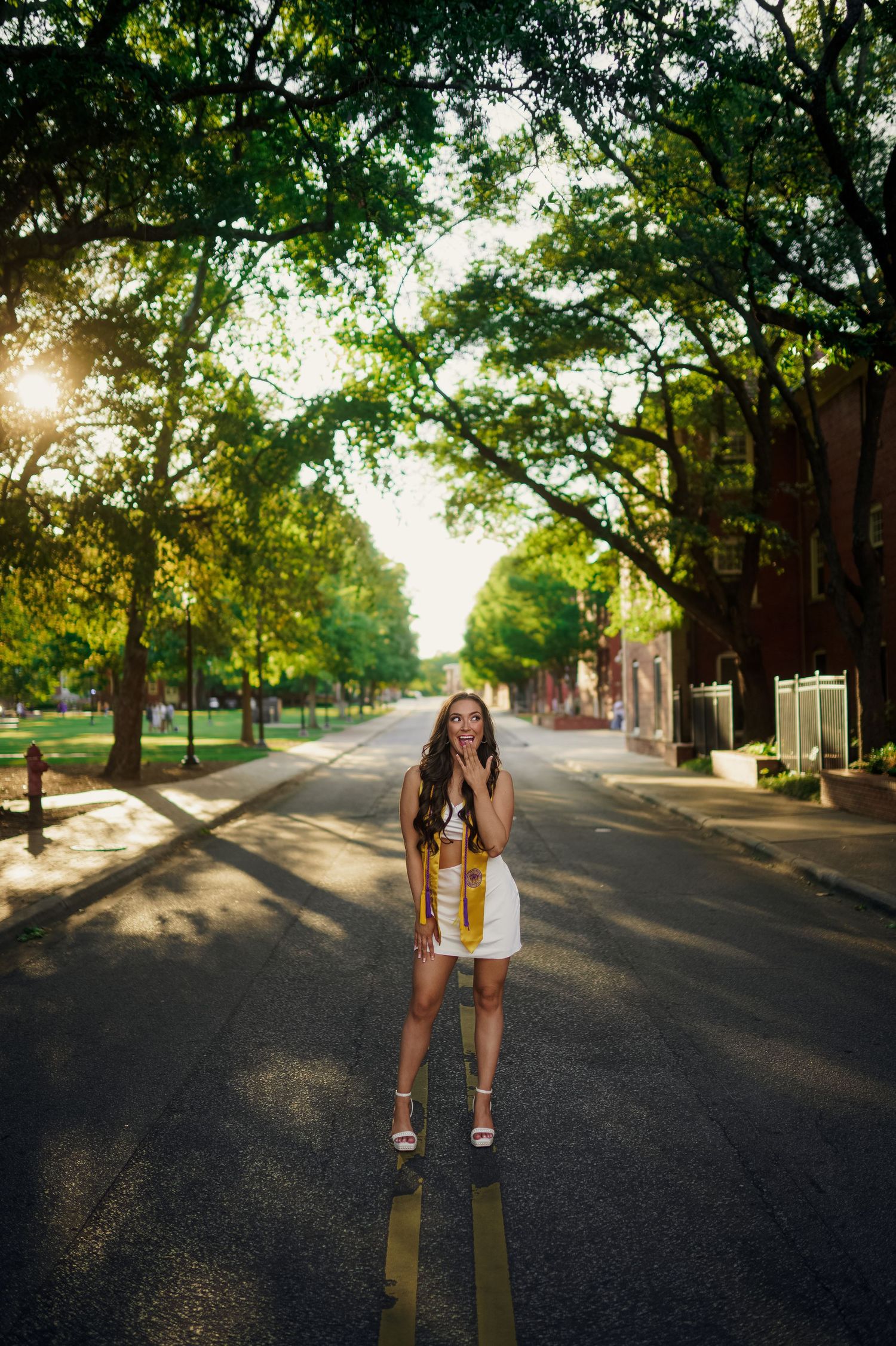 A ECU Graduate stands in the middle of a tree-lined street at golden hour with sunlight filtering through branches for grad photos.