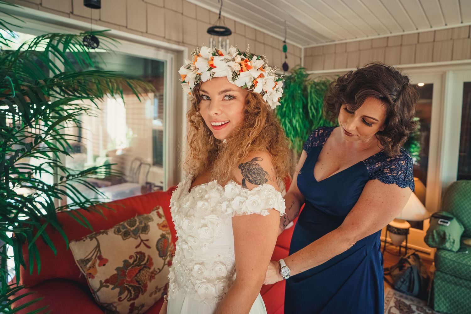 Getting ready scene with bride wearing flower crown and lace dress being helped with final touches.