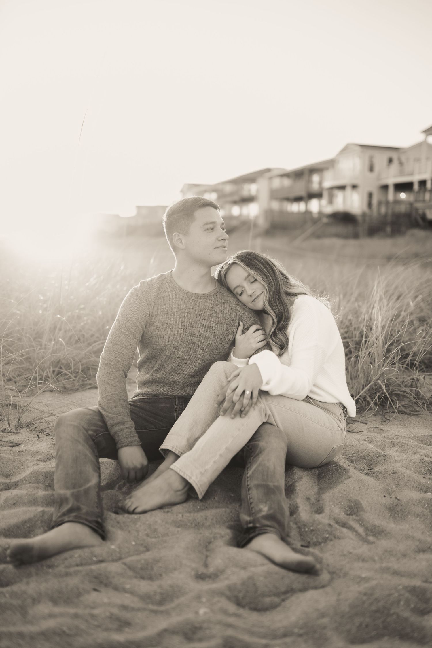 Black and white photo of a couple sitting on beach sand with houses in background.