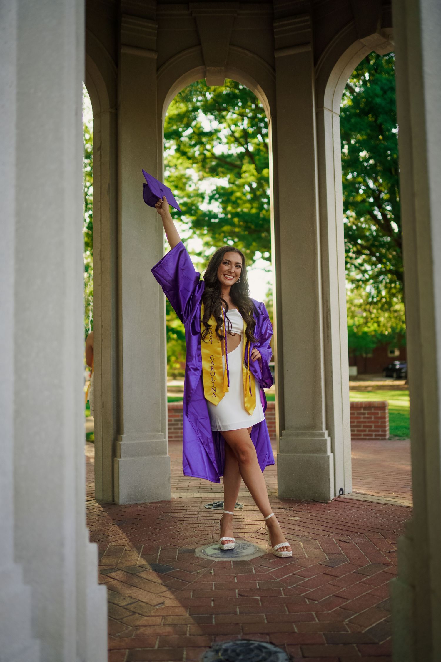 Graduate in purple cap and gown poses under ECU cupola on brick paved walkway during outdoor celebration Joan Avilez photography session.