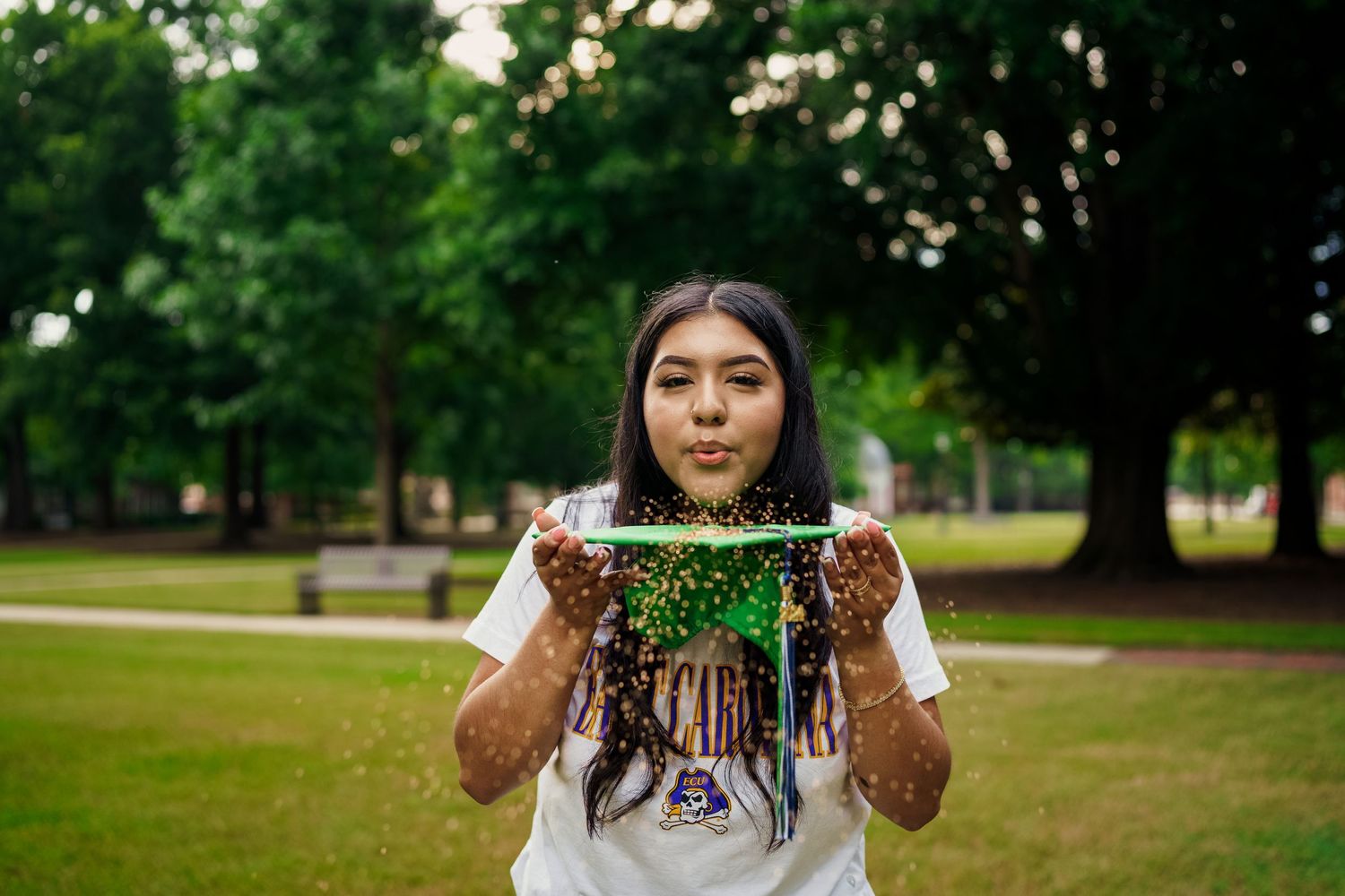 A decorative graduation tassel with dangling colorful threads is held up against a blurred green park background.