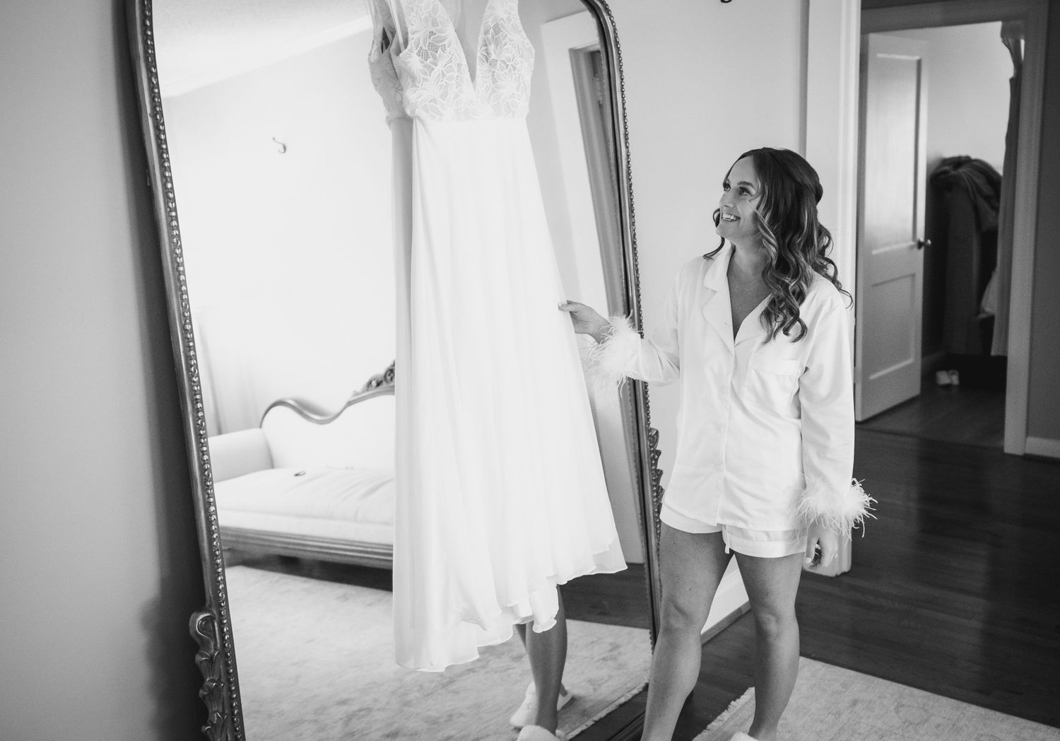 A wedding dress hangs in a sunlit room as someone in a white robe looks up at it.