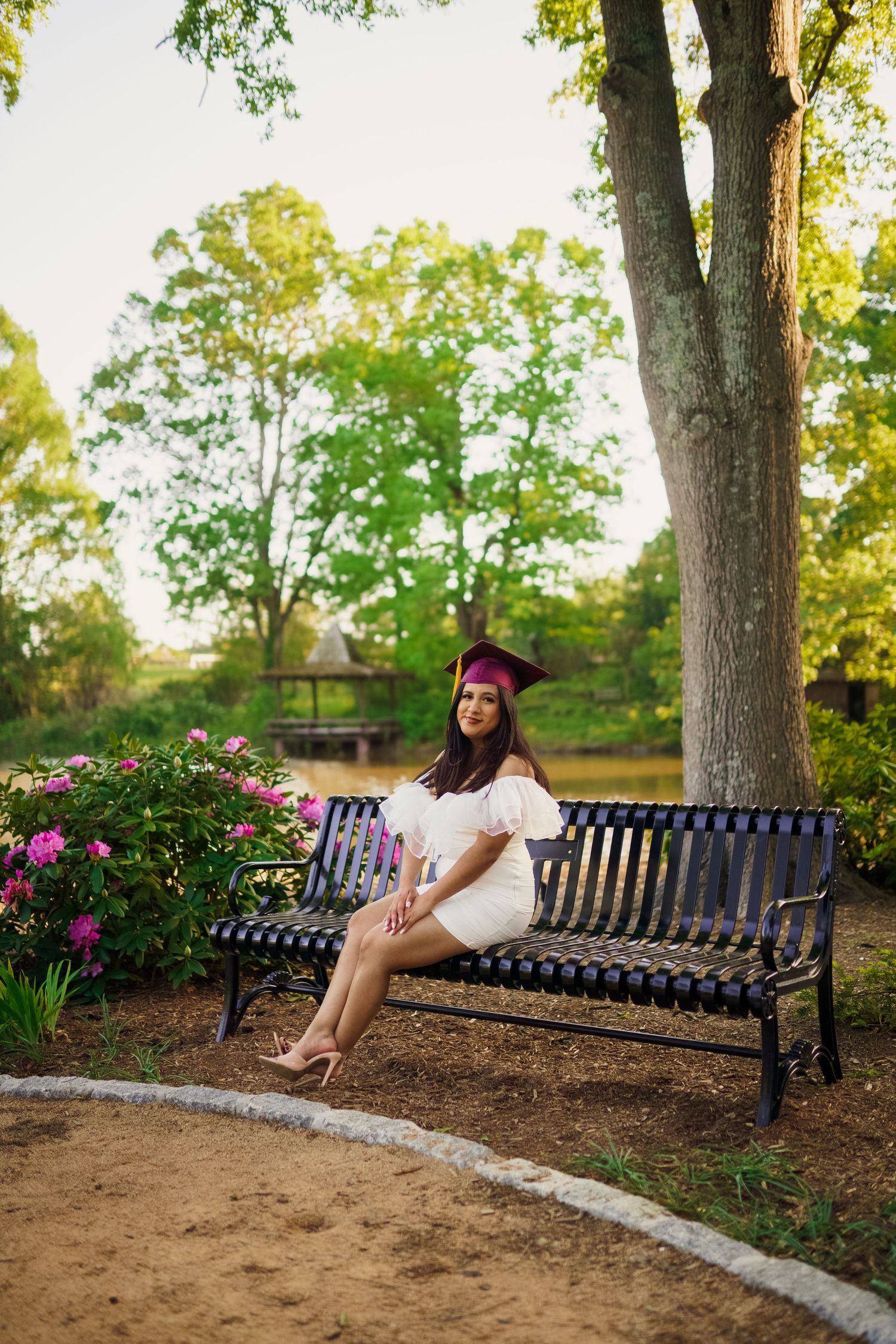 Graduate in cap and gown sitting on park bench surrounded by flowering bushes and trees.
