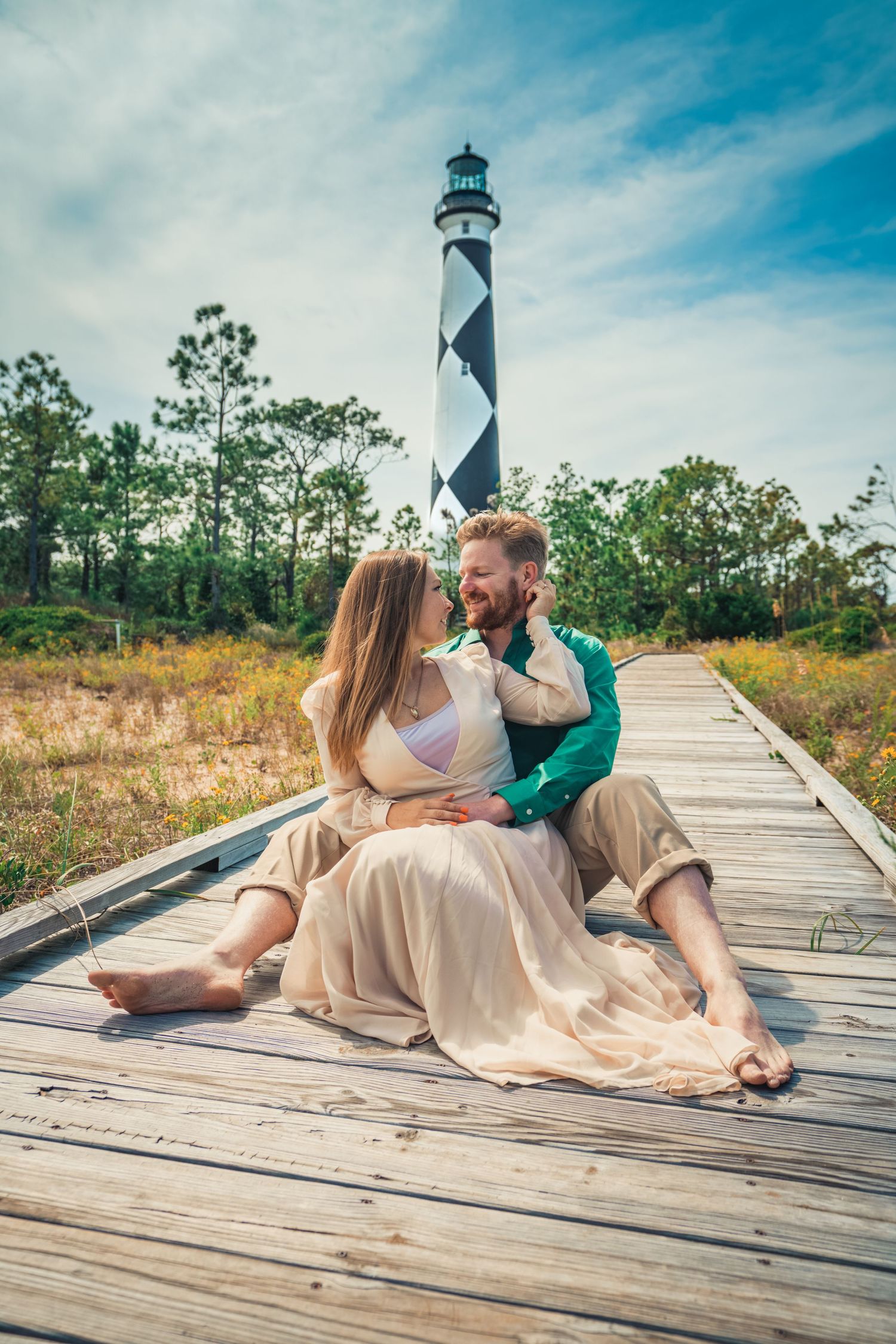 A couple sits intimately on a wooden boardwalk with a striped lighthouse towering behind them.