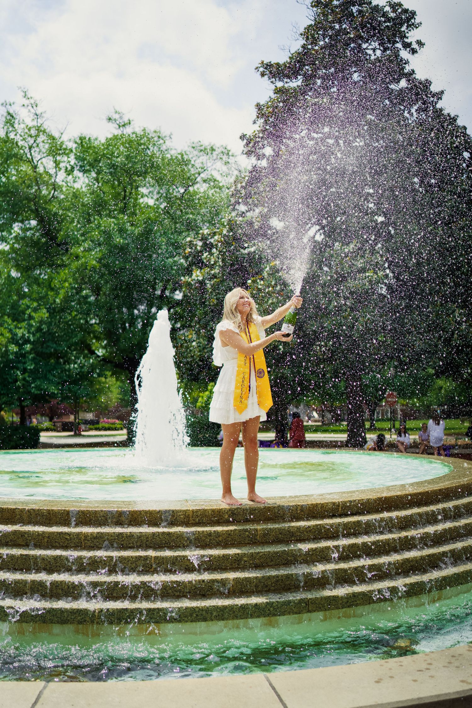 ECU Graduate poses on East carolina university's campus for the best ECU grad photos by Joan Avilez Photography. champagne spray at fountain