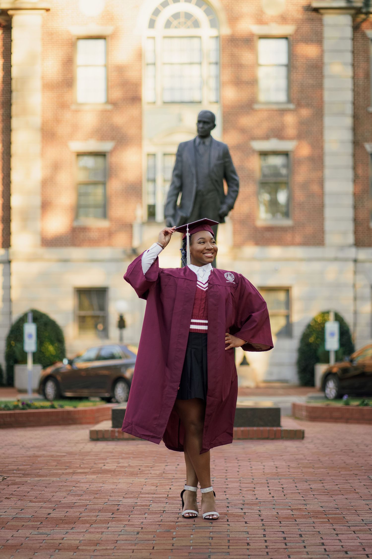 Graduate in maroon academic regalia poses in front of historic brick building with arched windows.