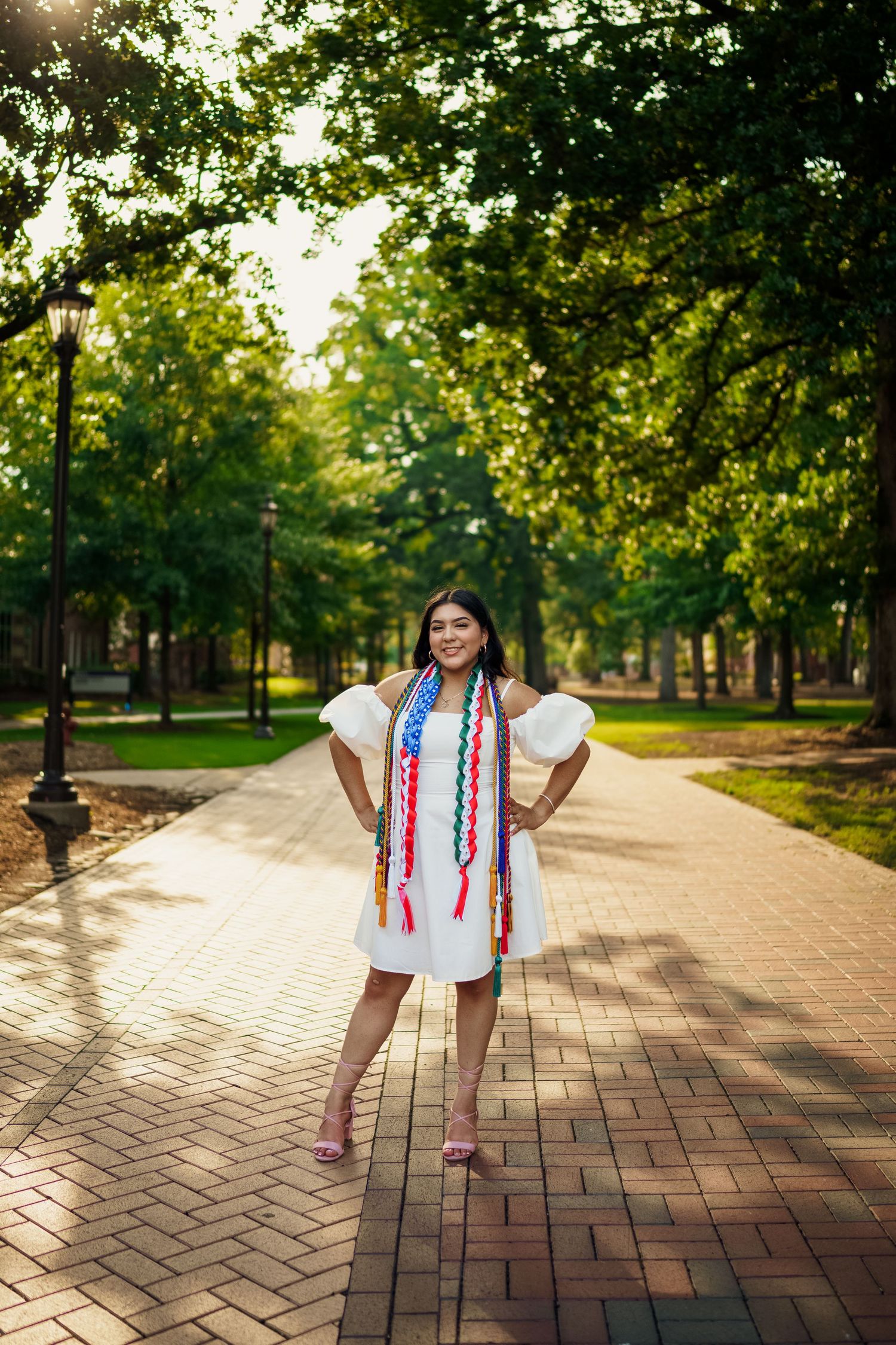 Graduate poses on brick pathway under green tree canopy wearing white dress and graduation cords.