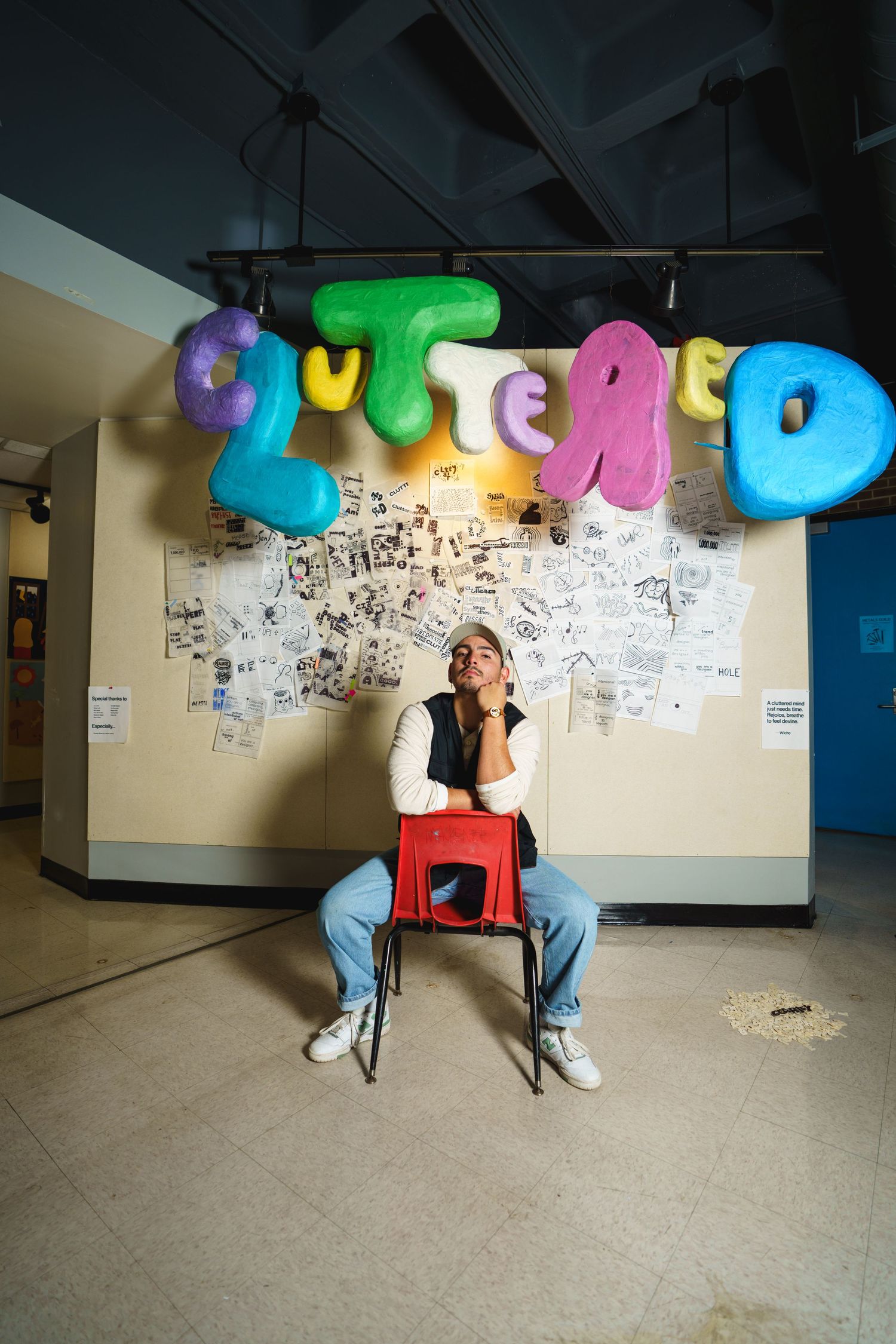 Someone sits on a red chair beneath colorful inflatable letters on a wall with sketches in a hallway.