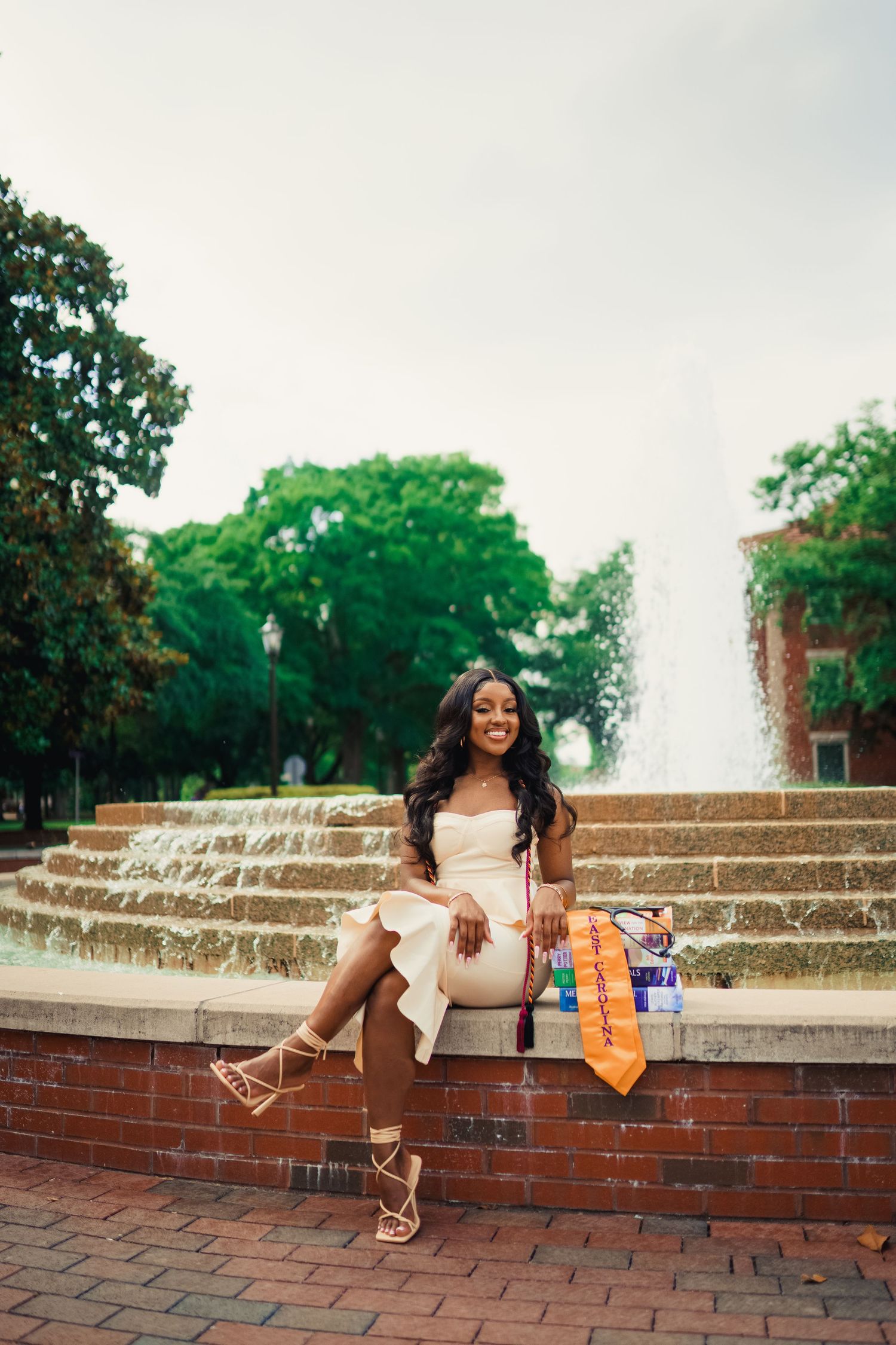 A graduate in a cream dress sits on ledge in front of a fountain wearing graduation regalia and posing with her nursing text books.