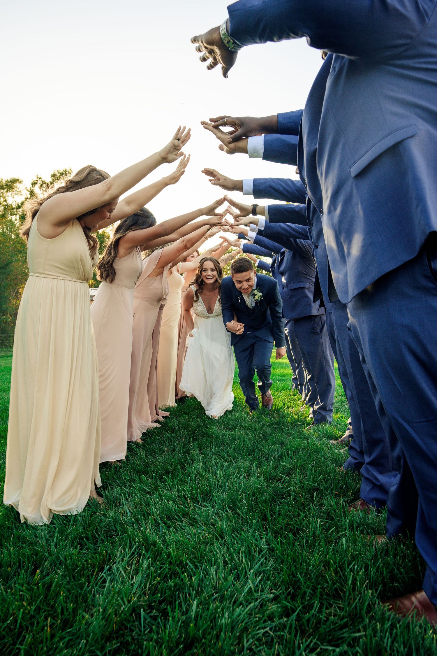 Wedding party forms tunnel on grass with bridesmaids in blush and groomsmen in navy.