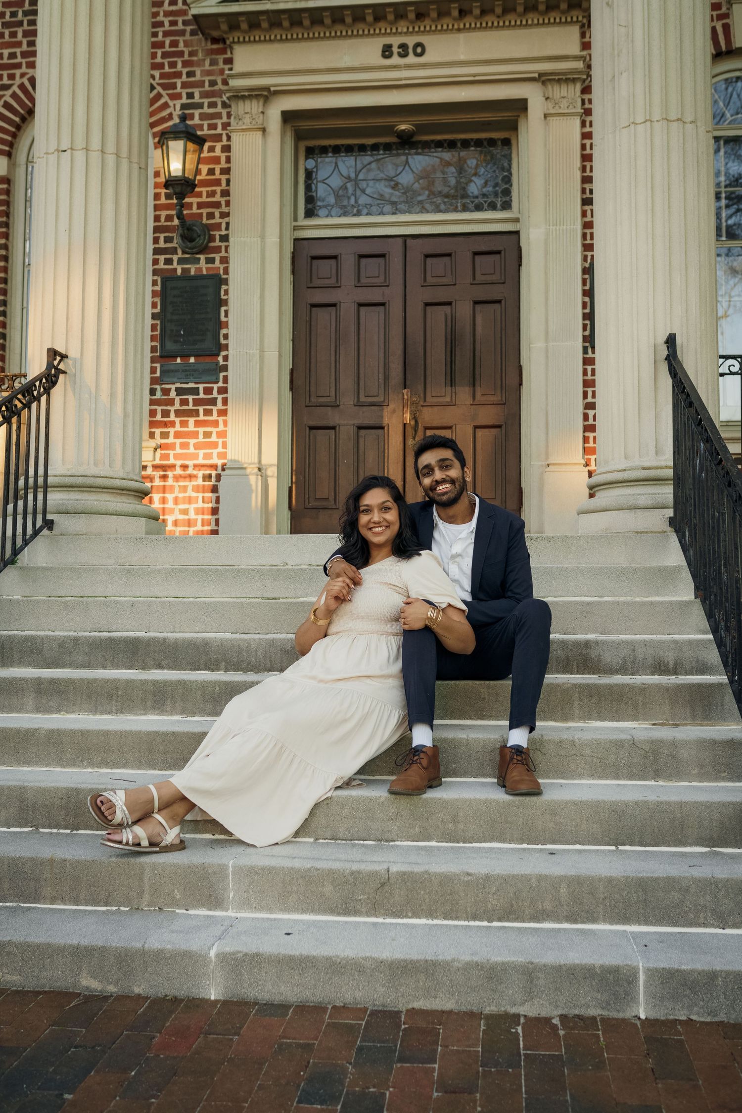 A couple sits casually on stone steps of a historic brick building with white columns.