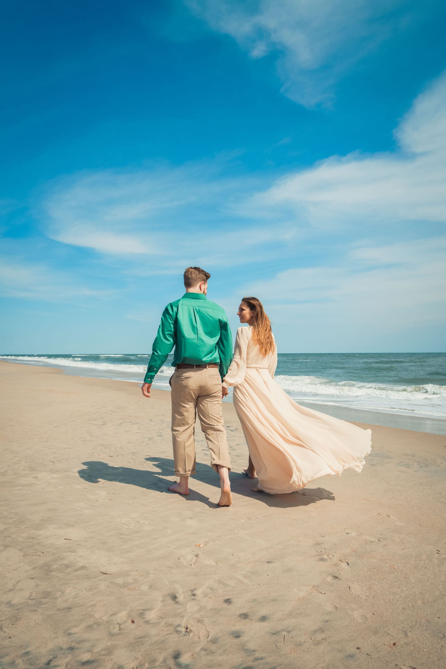 A couple walks along a beach shoreline with flowing dress and green jacket against blue sky.