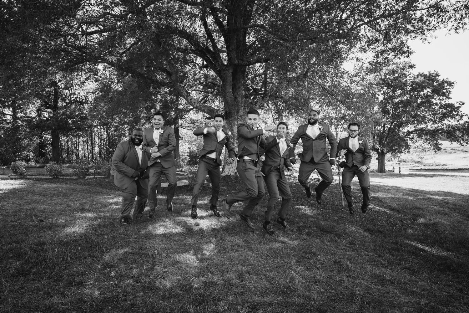Black and white photo of groomsmen posing casually outdoors under large tree.