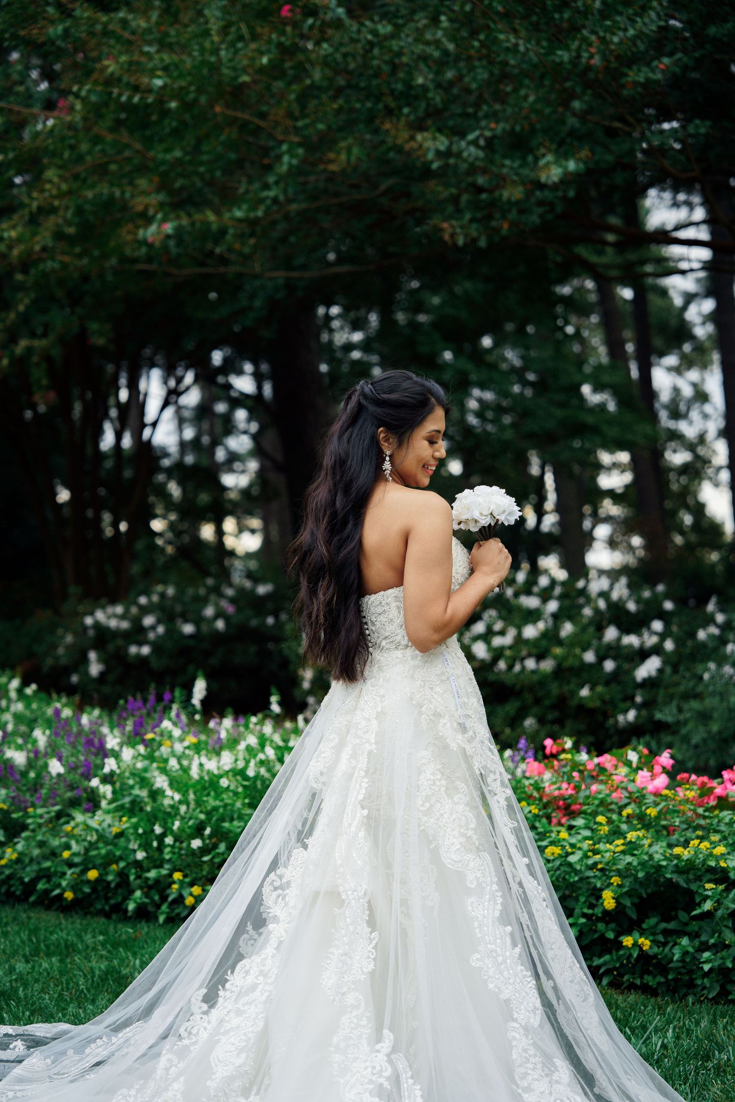 Bride in white lace gown with long train holds bouquet while posing in garden with colorful flowers.