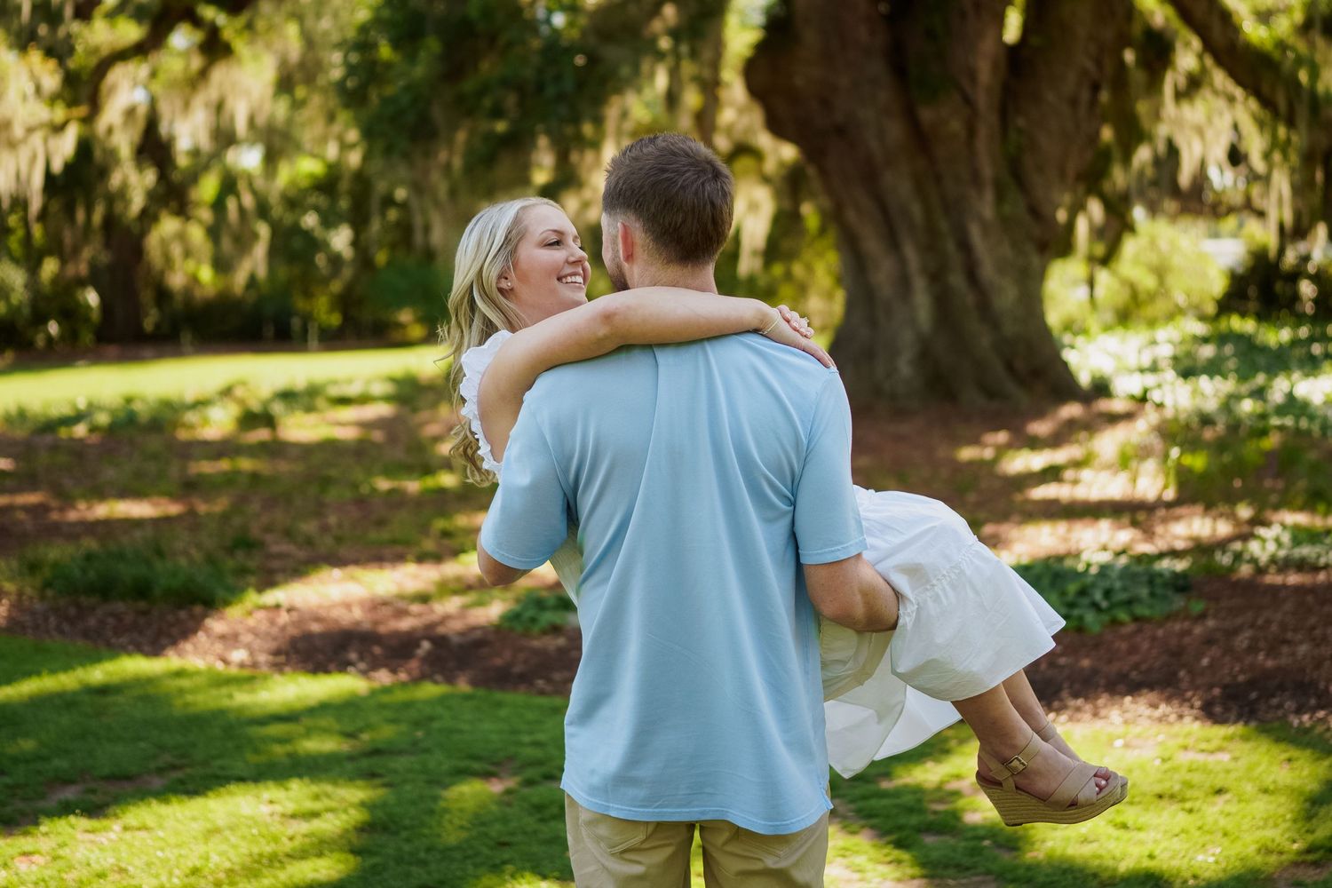 A couple embraces in a sunlit garden with Spanish moss draped trees in the background.