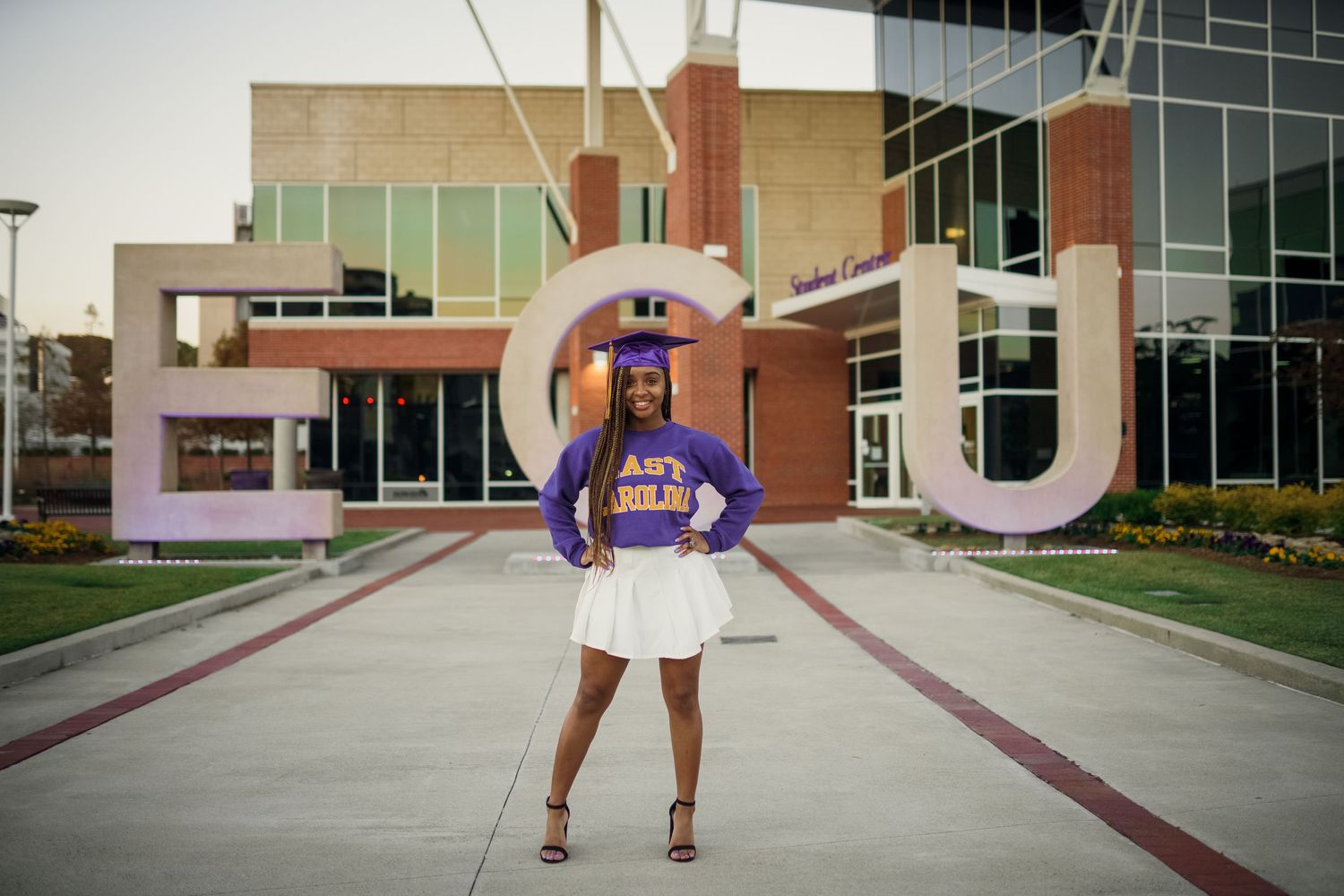East Carolina university student wearing a purple ECU shirt. stands in front of a ECU student center for grad photos by Joan Avilez