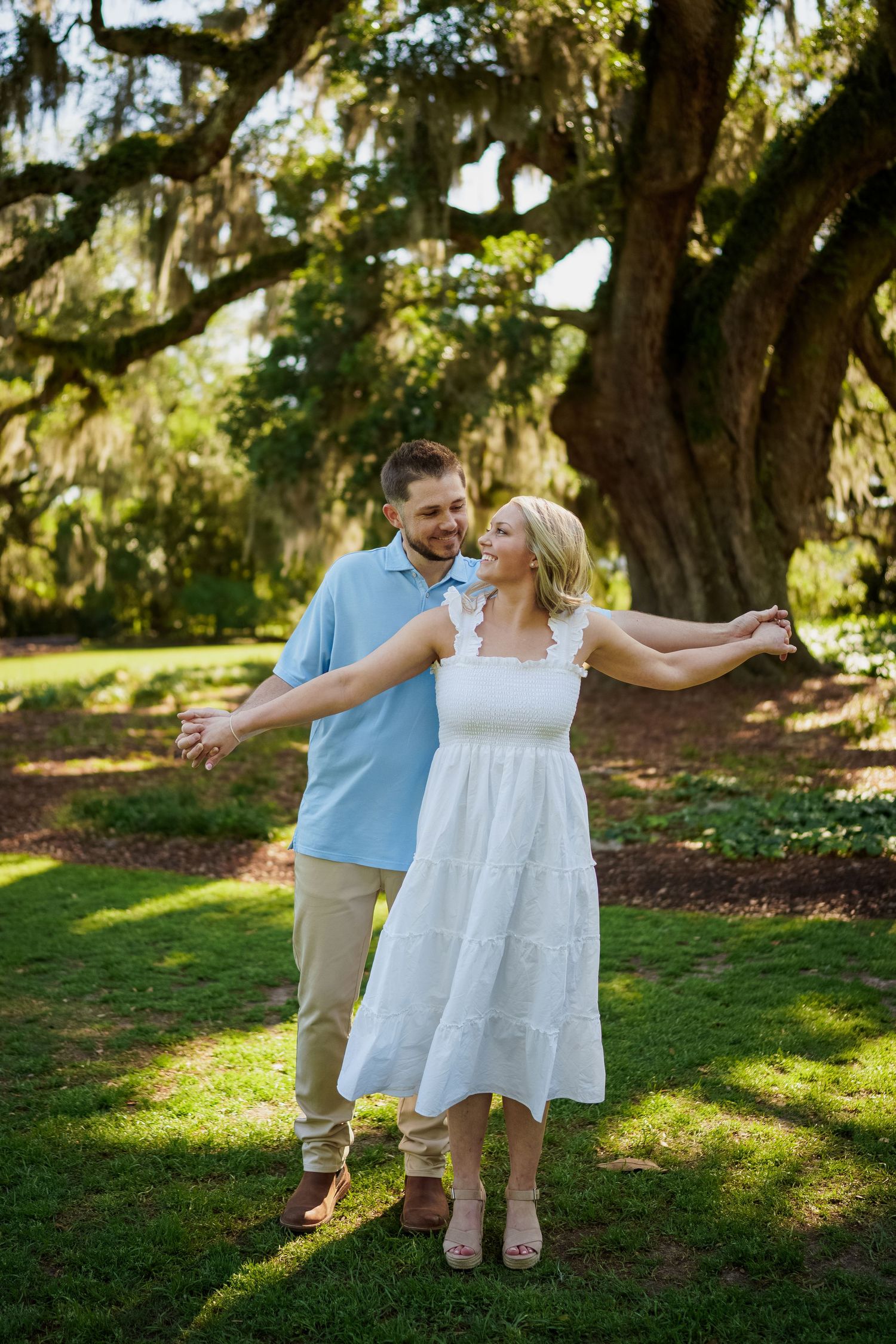 A joyful couple dances together on a grassy lawn beneath moss-covered trees.