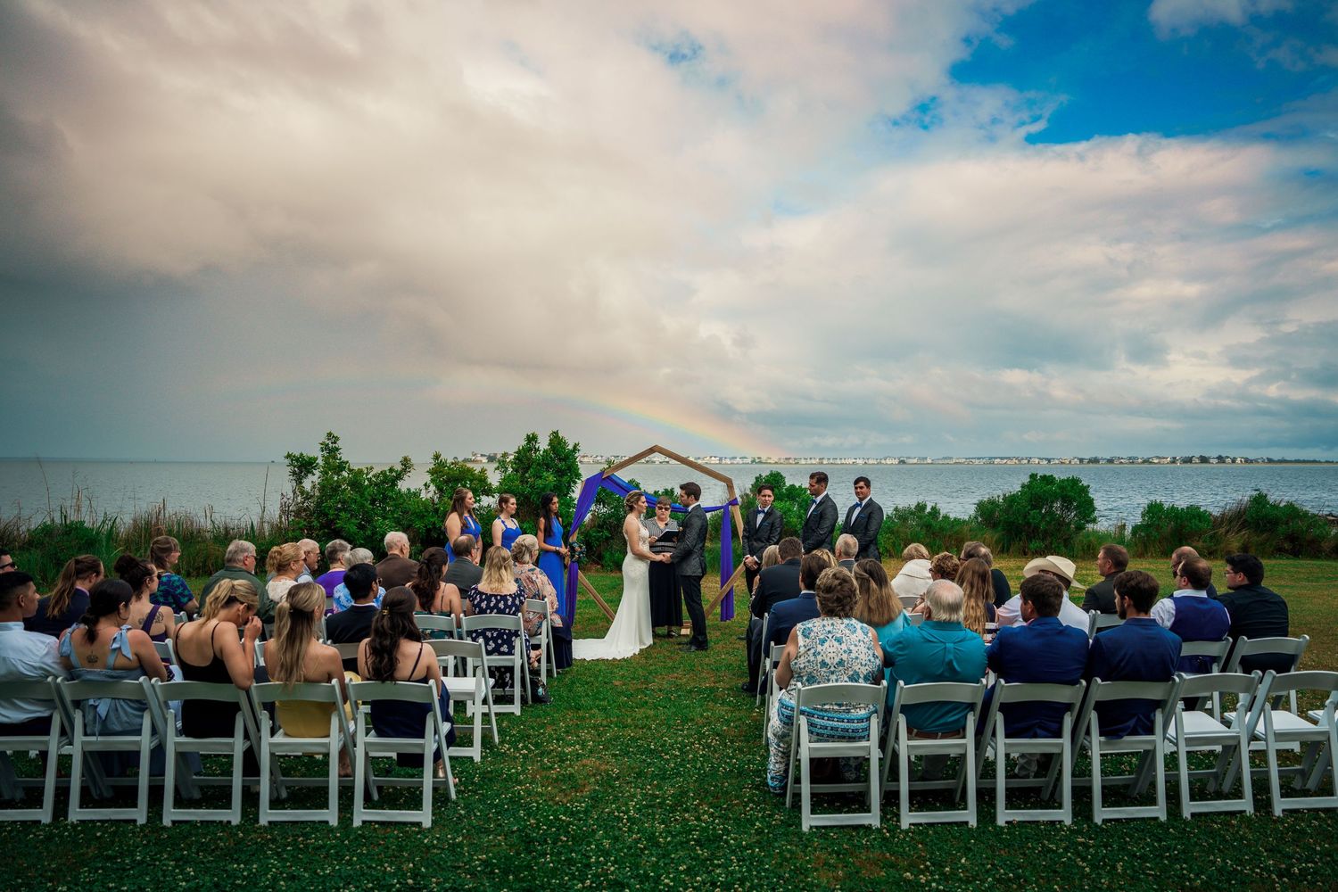 Outdoor wedding ceremony setup with white chairs on grass overlooking water under cloudy sky.