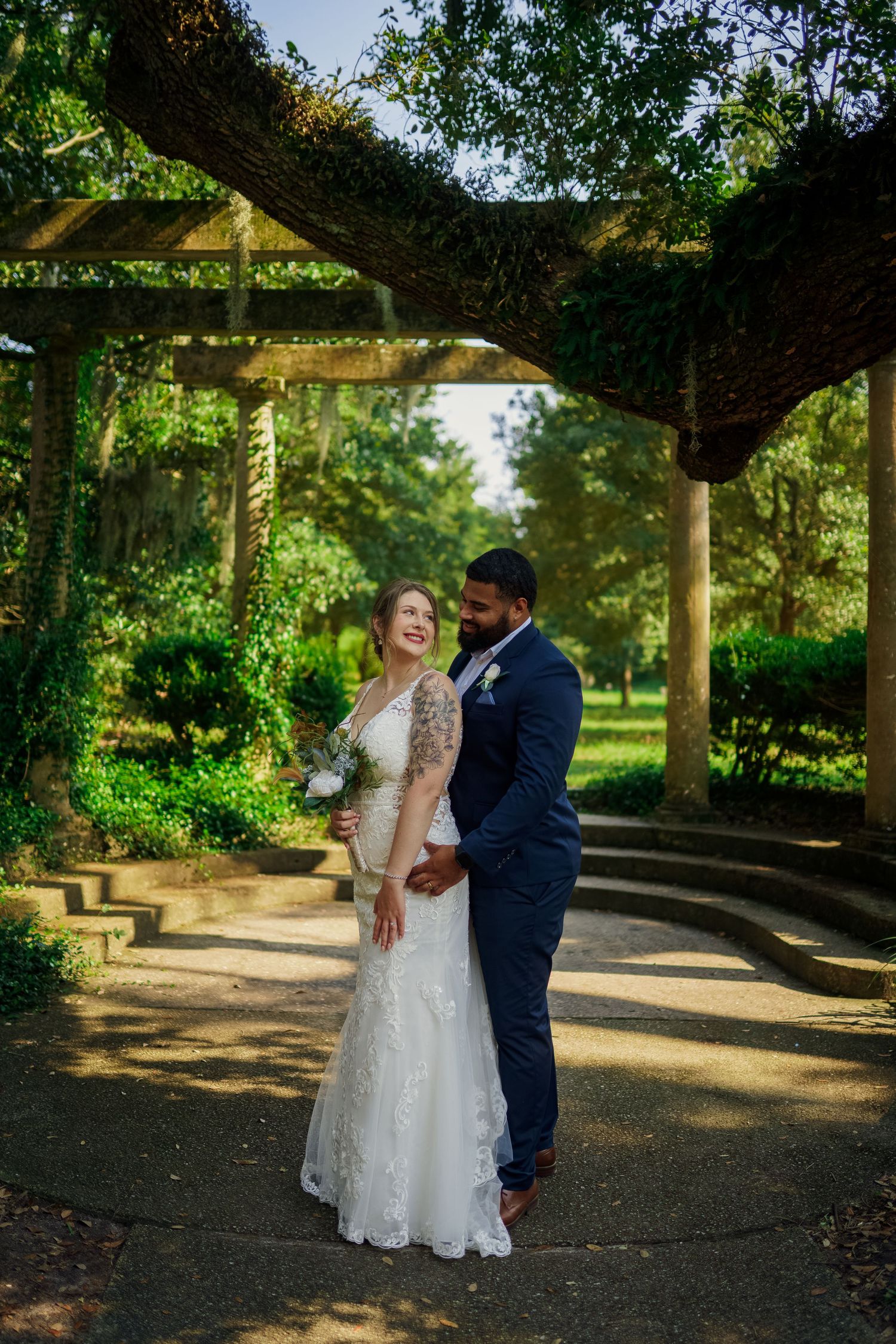 Newlyweds share intimate moment under vine-covered garden pergola in natural lighting.