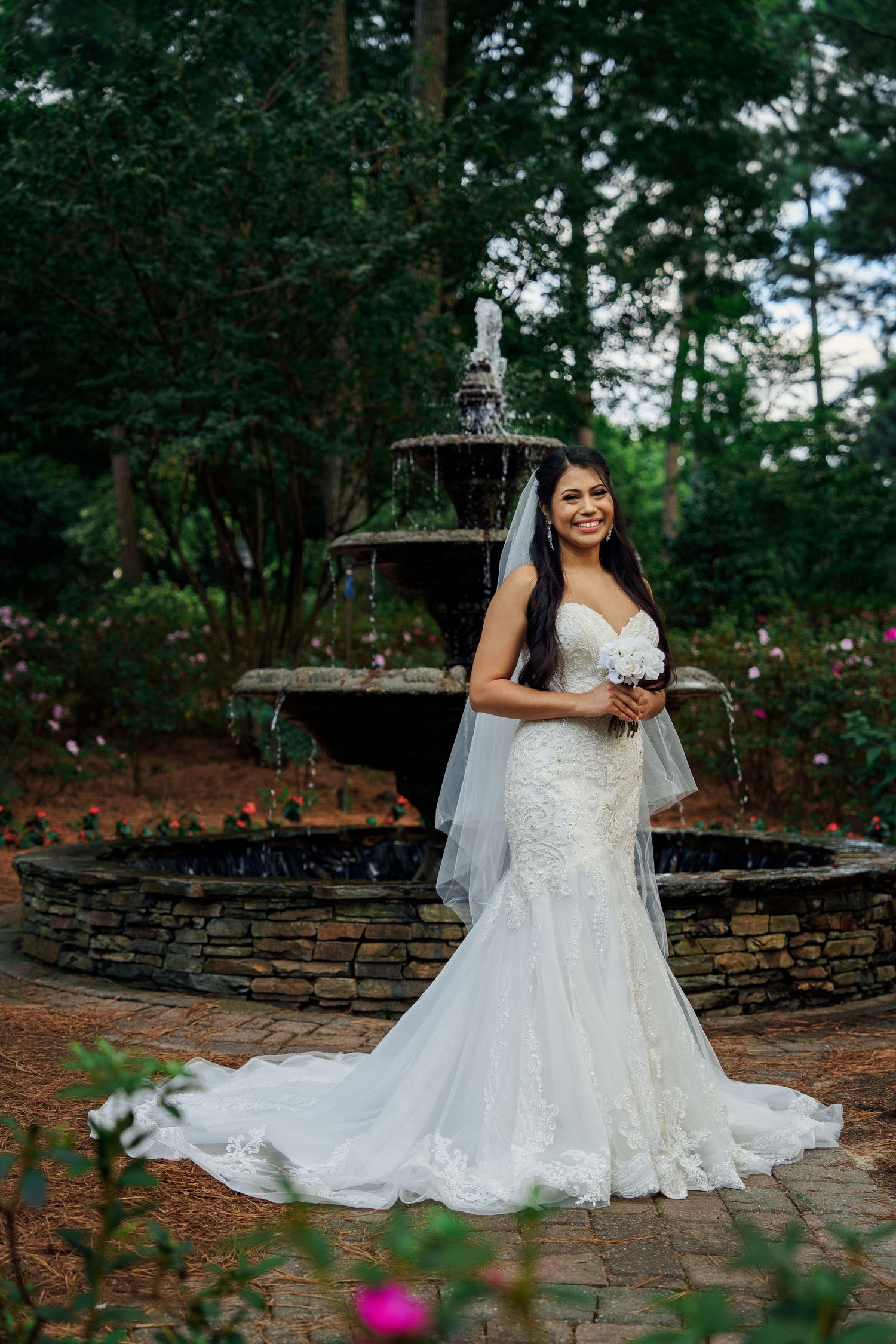 Elegant bridal portrait in a white lace gown standing by a stone fountain in a garden setting.