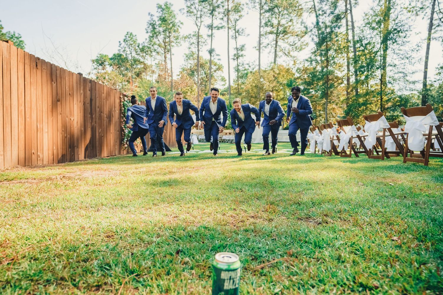 Wedding party in navy blue attire walking together on grass with wooden fence in background.