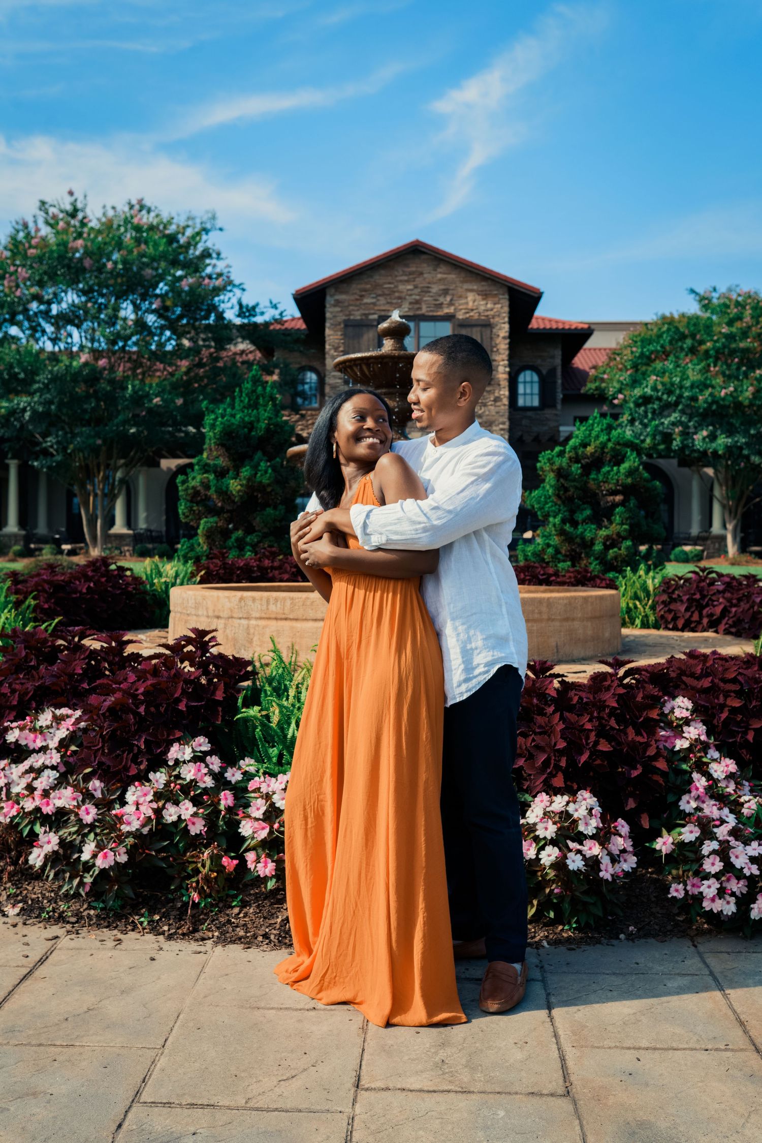 A couple embraces in front of a stone building with pink flowers and landscaping in the foreground.