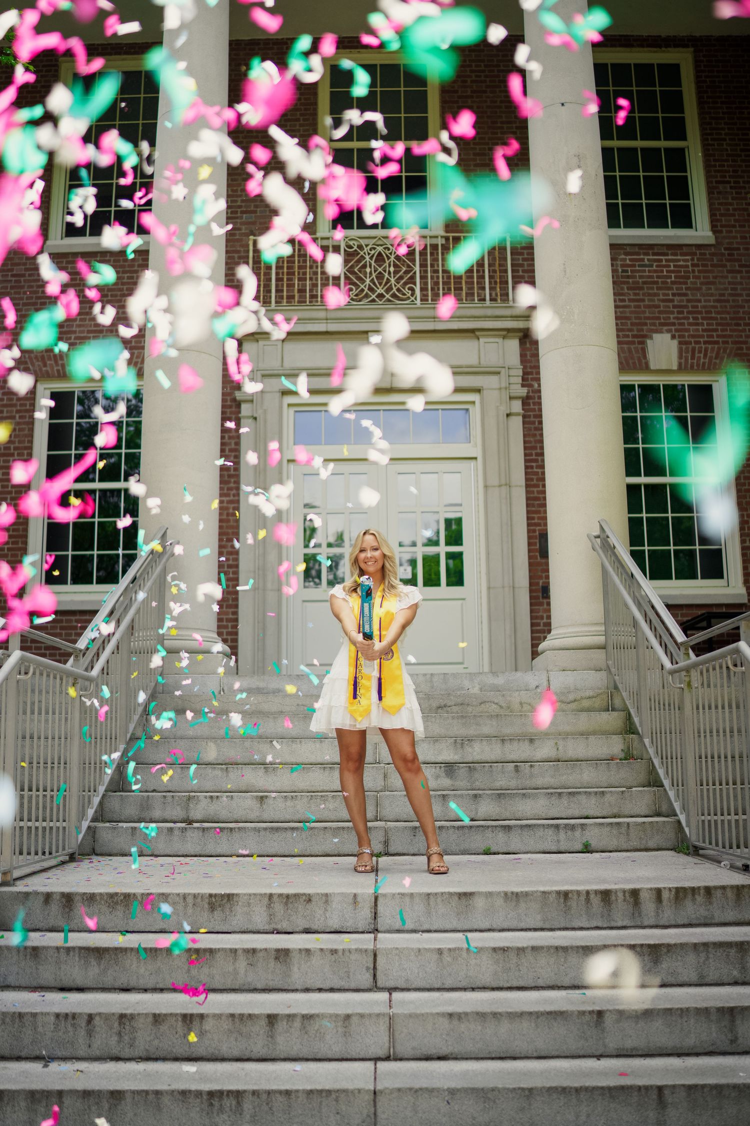 Celebratory scene on grand stone steps with pink and turquoise confetti floating around figure in white graduation dress for grad photos