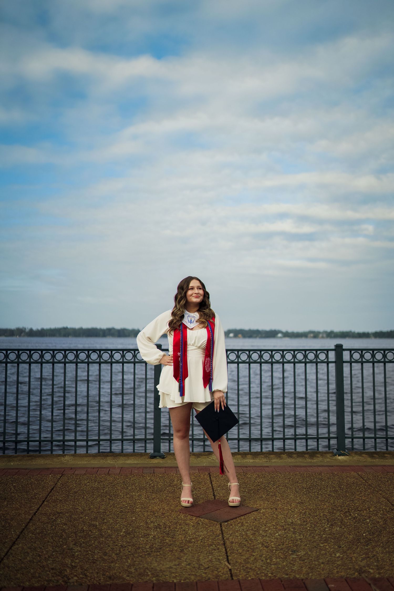 Graduate in white dress poses along waterfront railing under cloudy sky.