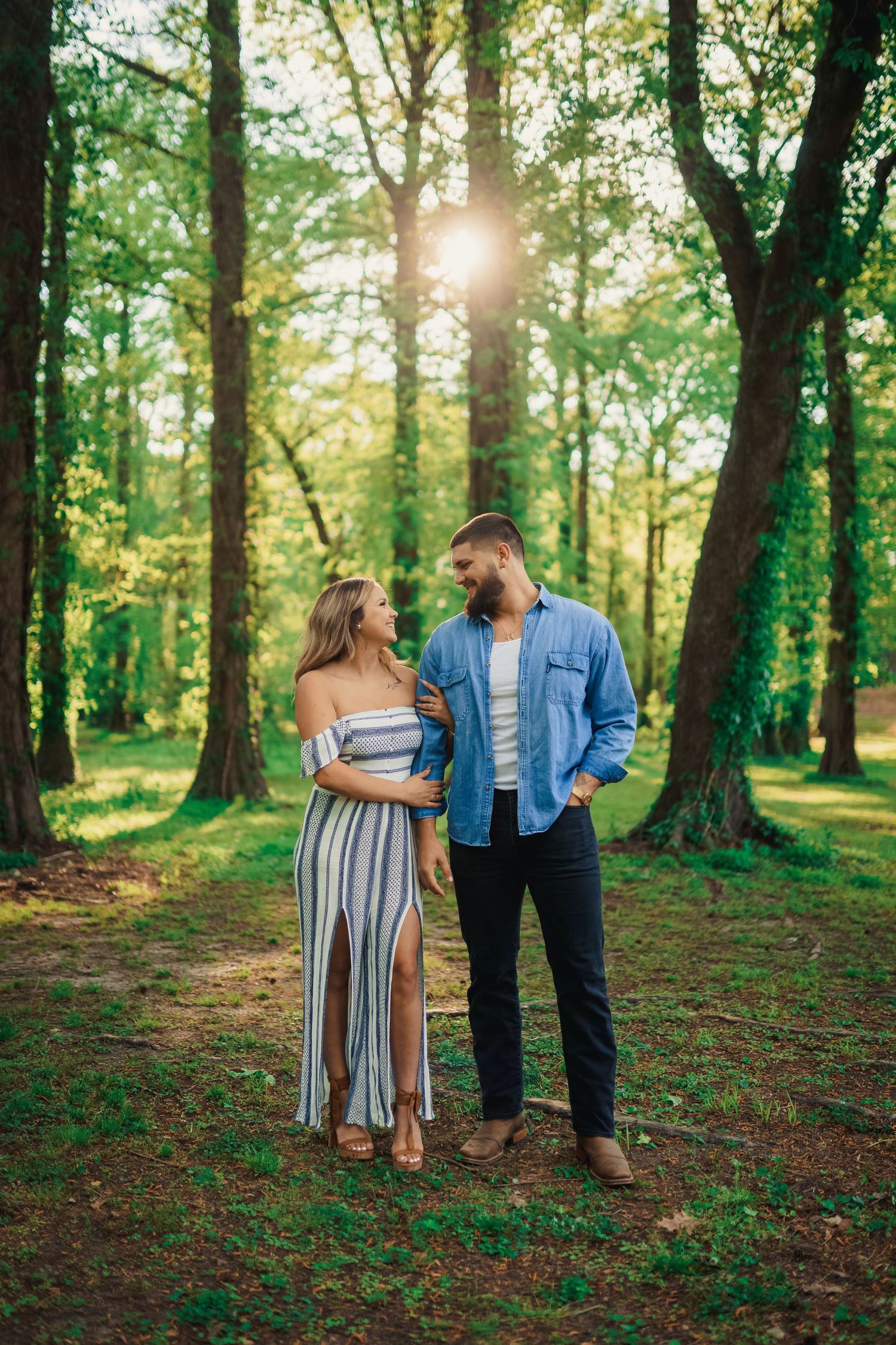 A couple in coordinated outfits stands together on a forest path dappled with sunlight.