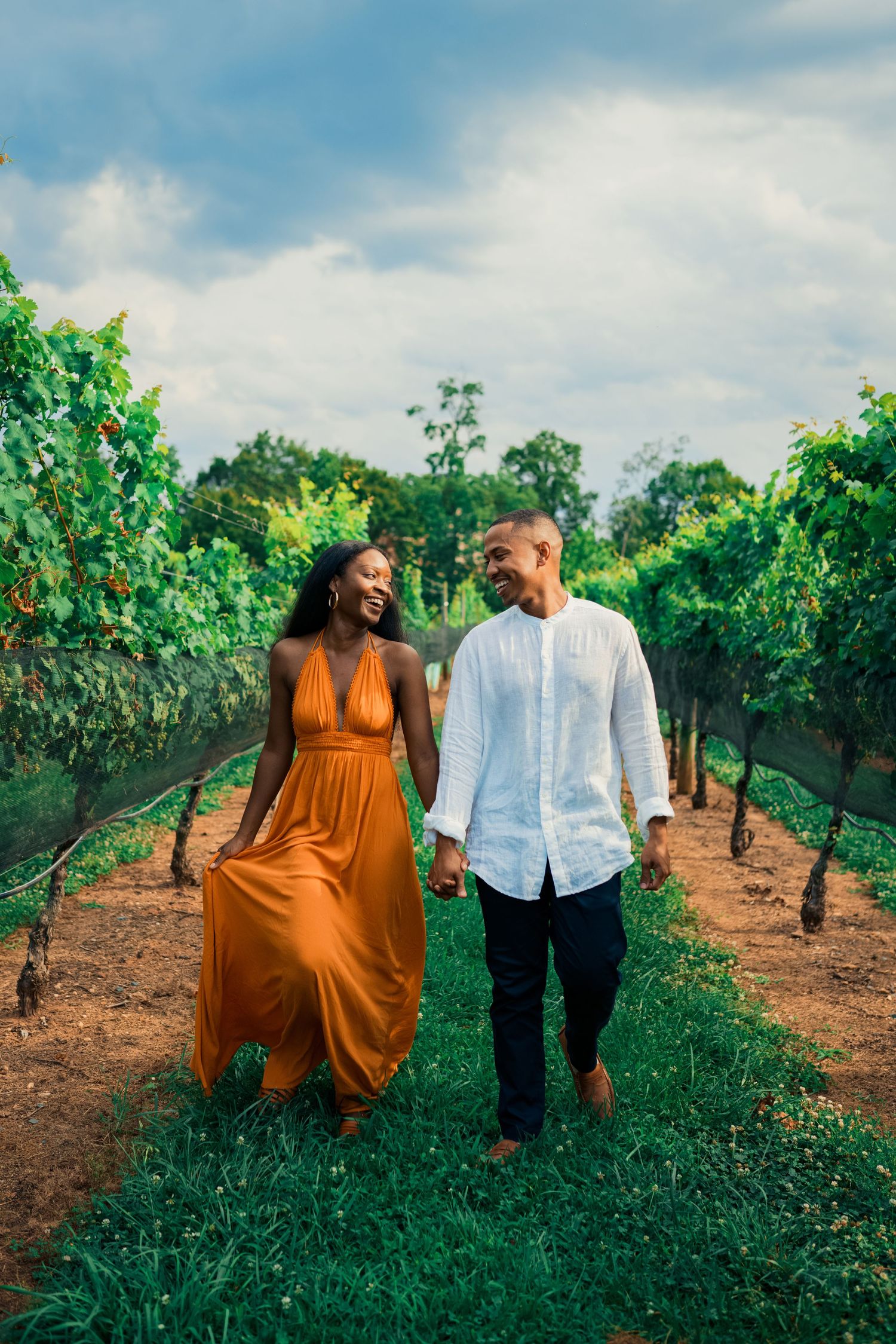 A couple walks hand in hand through a lush vineyard with one wearing an orange dress and the other in white.