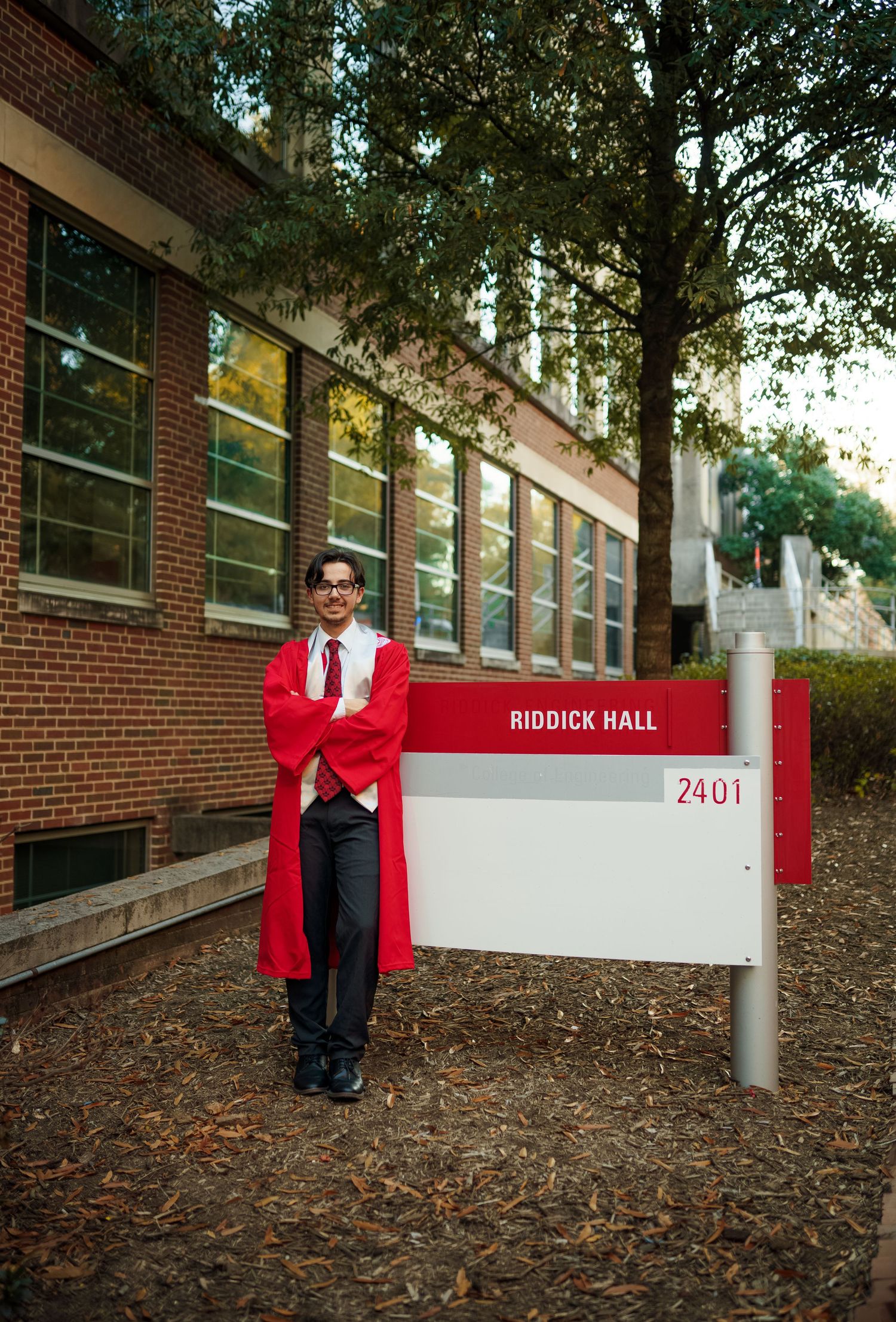 Graduate stands in red academic regalia next to Riddick Hall building sign on brick campus building.