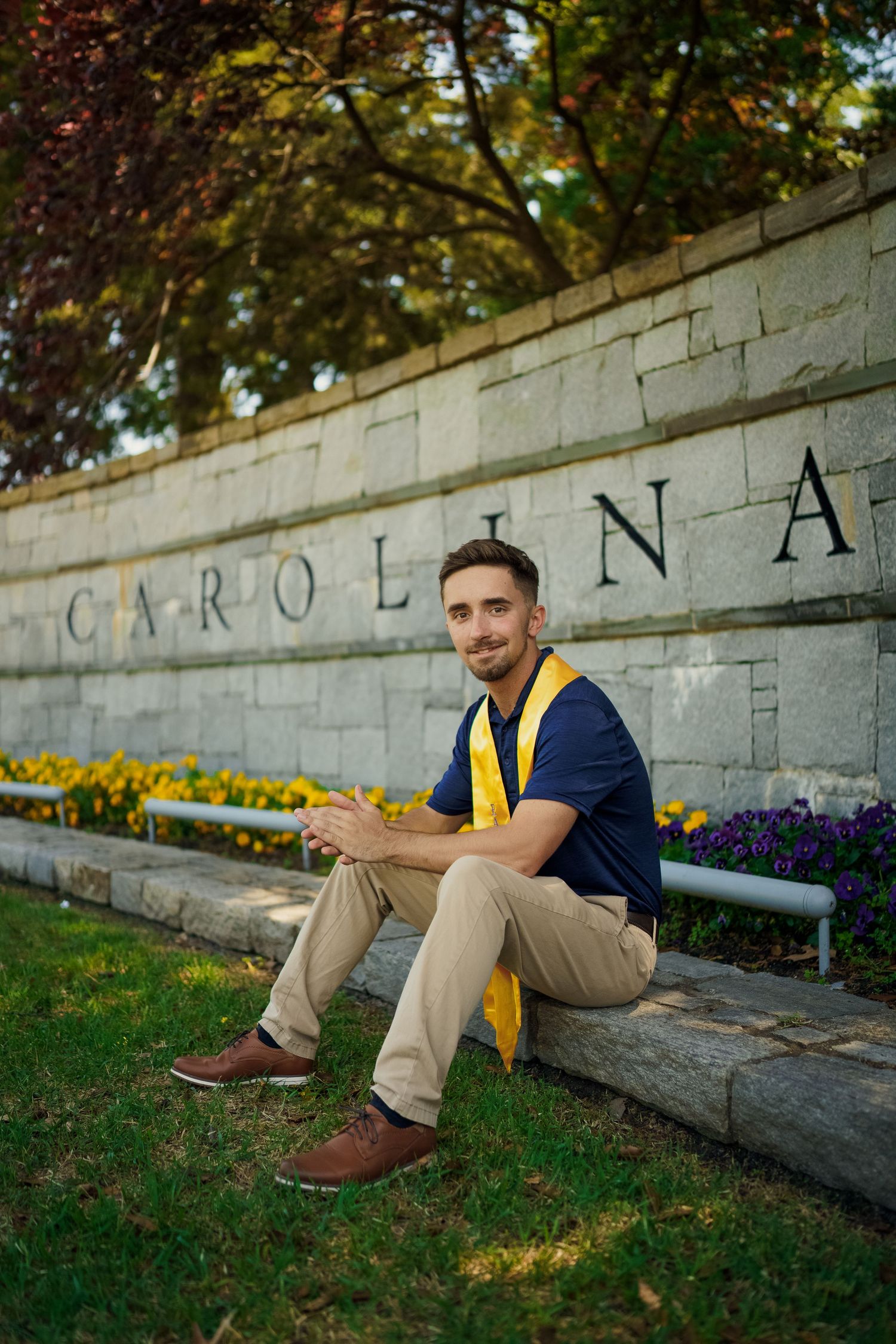 Collegiate scene at East Carolina with someone seated on stone wall wearing yellow stole and navy shirt near blooming flowers for graduation