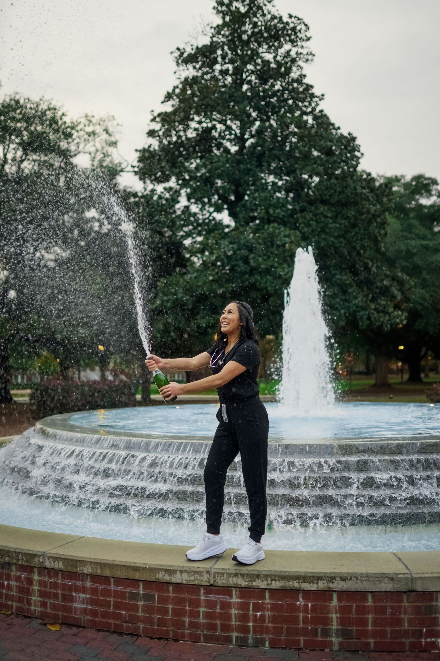 A person in black stands by a fountain spraying water in a scenic outdoor setting.