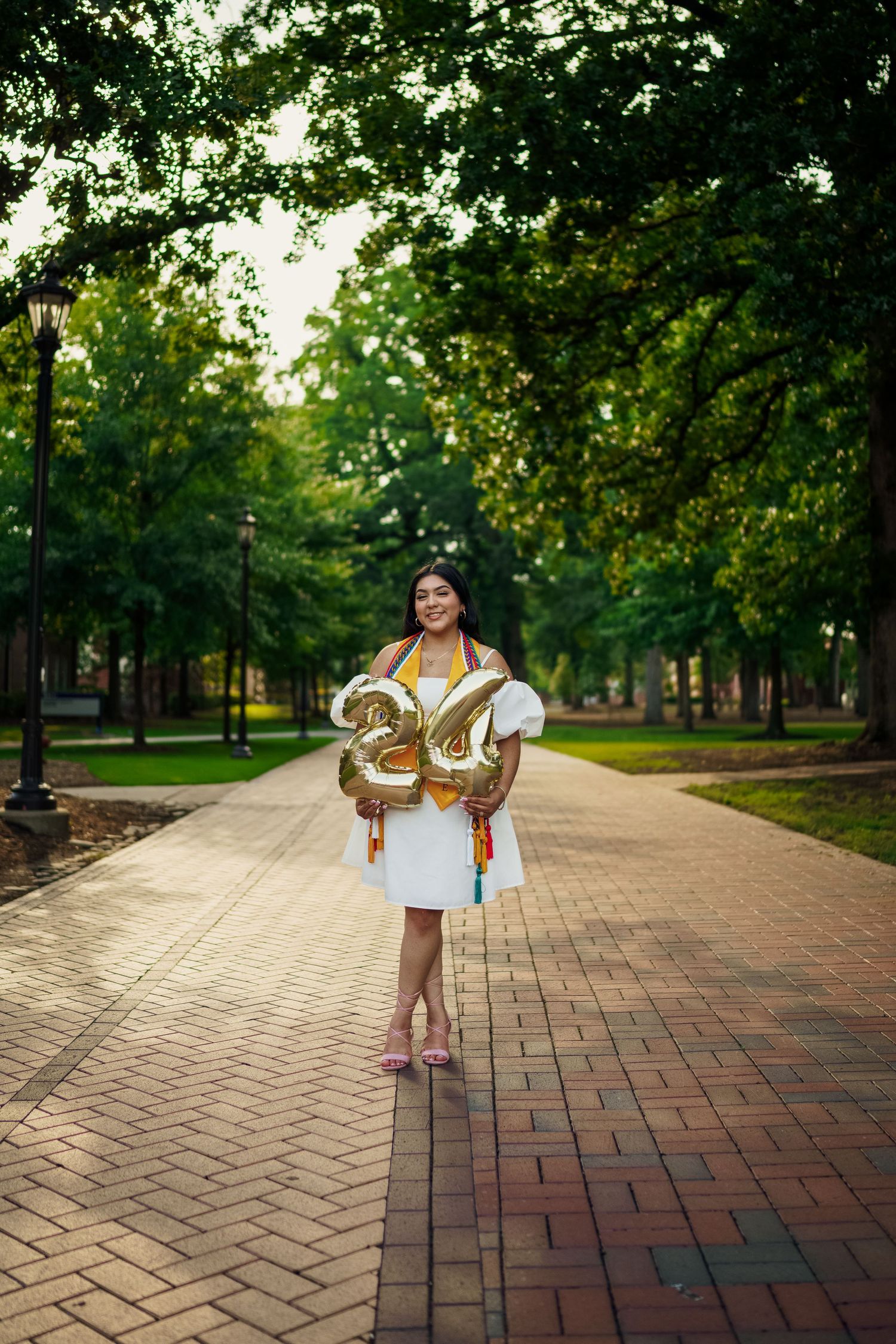 Graduate holds gold number balloons on tree-lined brick pathway in white dress.