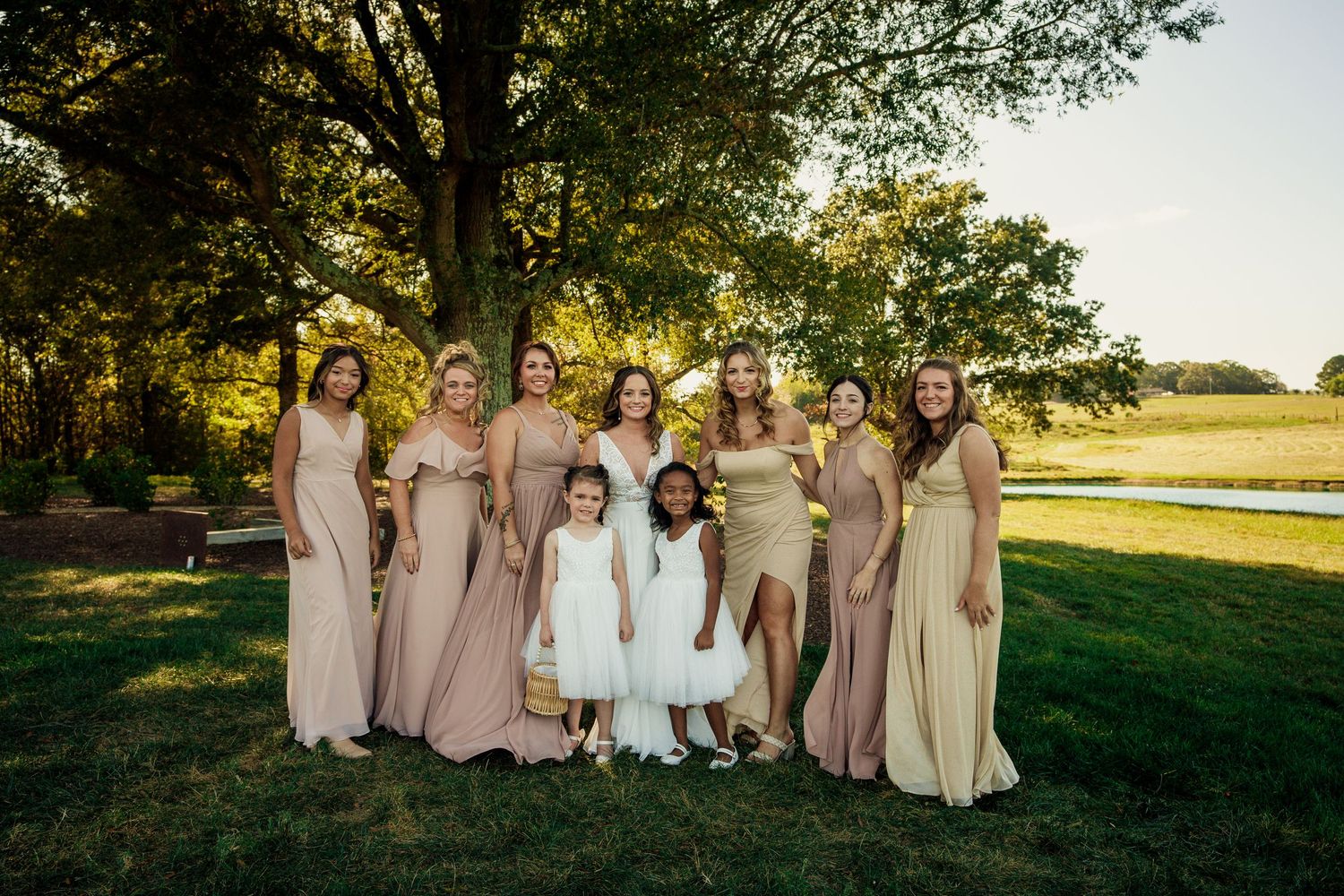 Bridesmaids in blush gowns pose with flower girls on grassy lawn at sunset.