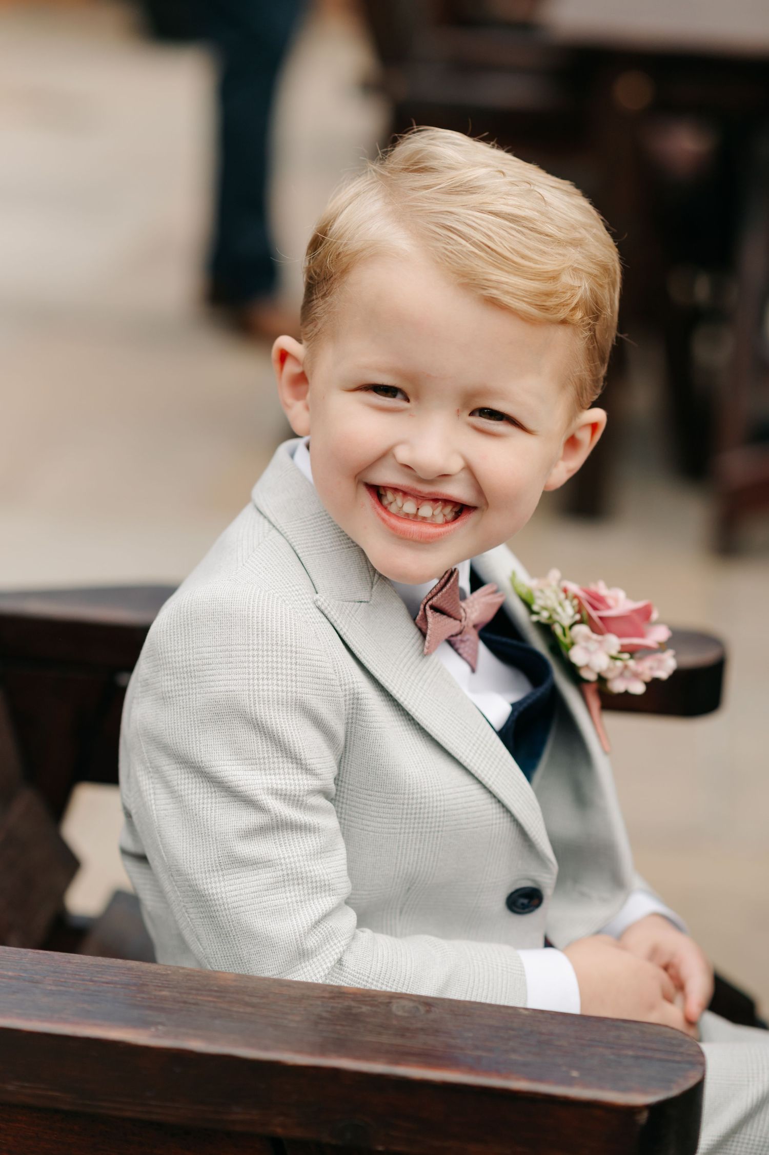 A young ring bearer in a light gray suit smiles while seated at a wedding reception.