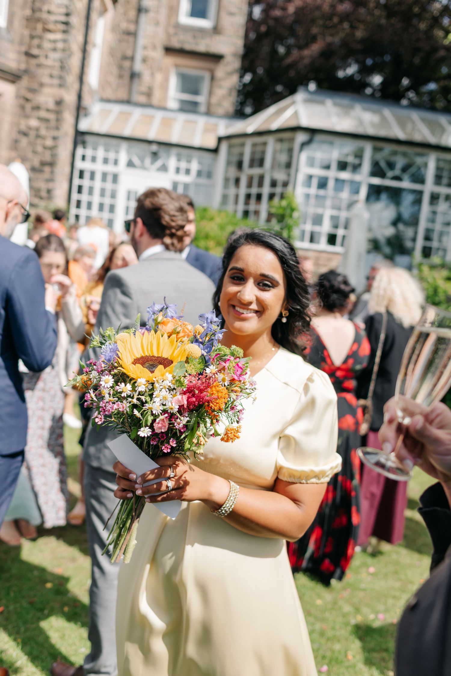 A person in cream-colored dress holds a bright bouquet featuring sunflowers and wildflowers.