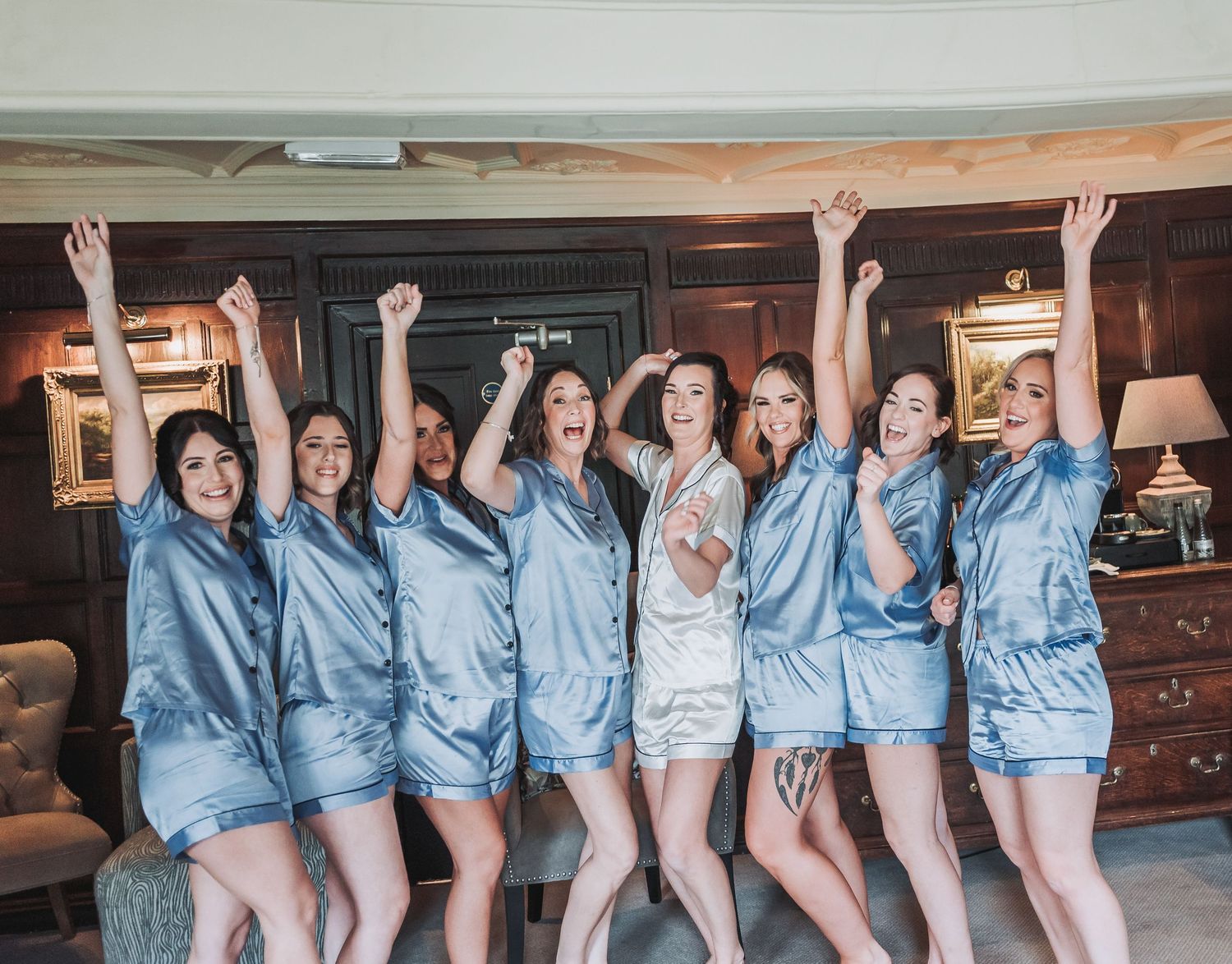 Bridesmaids in matching blue pajamas celebrating with raised arms in a hotel room.