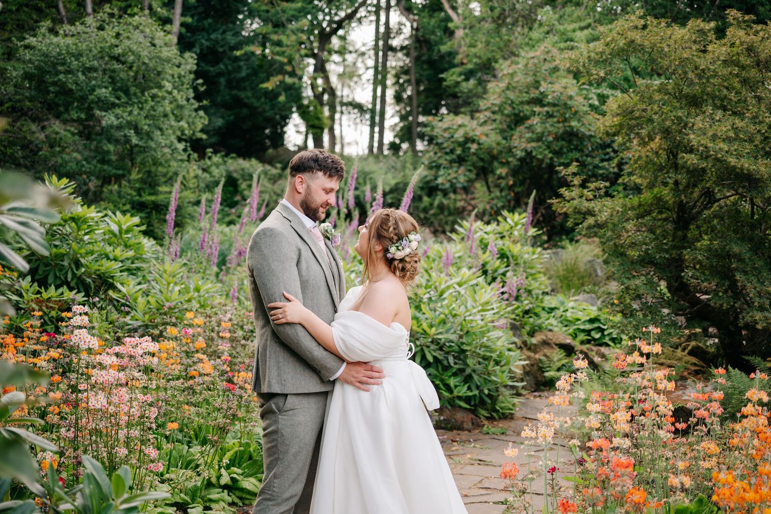 A couple shares a romantic moment in a lush garden path surrounded by vibrant orange flowers and greenery.