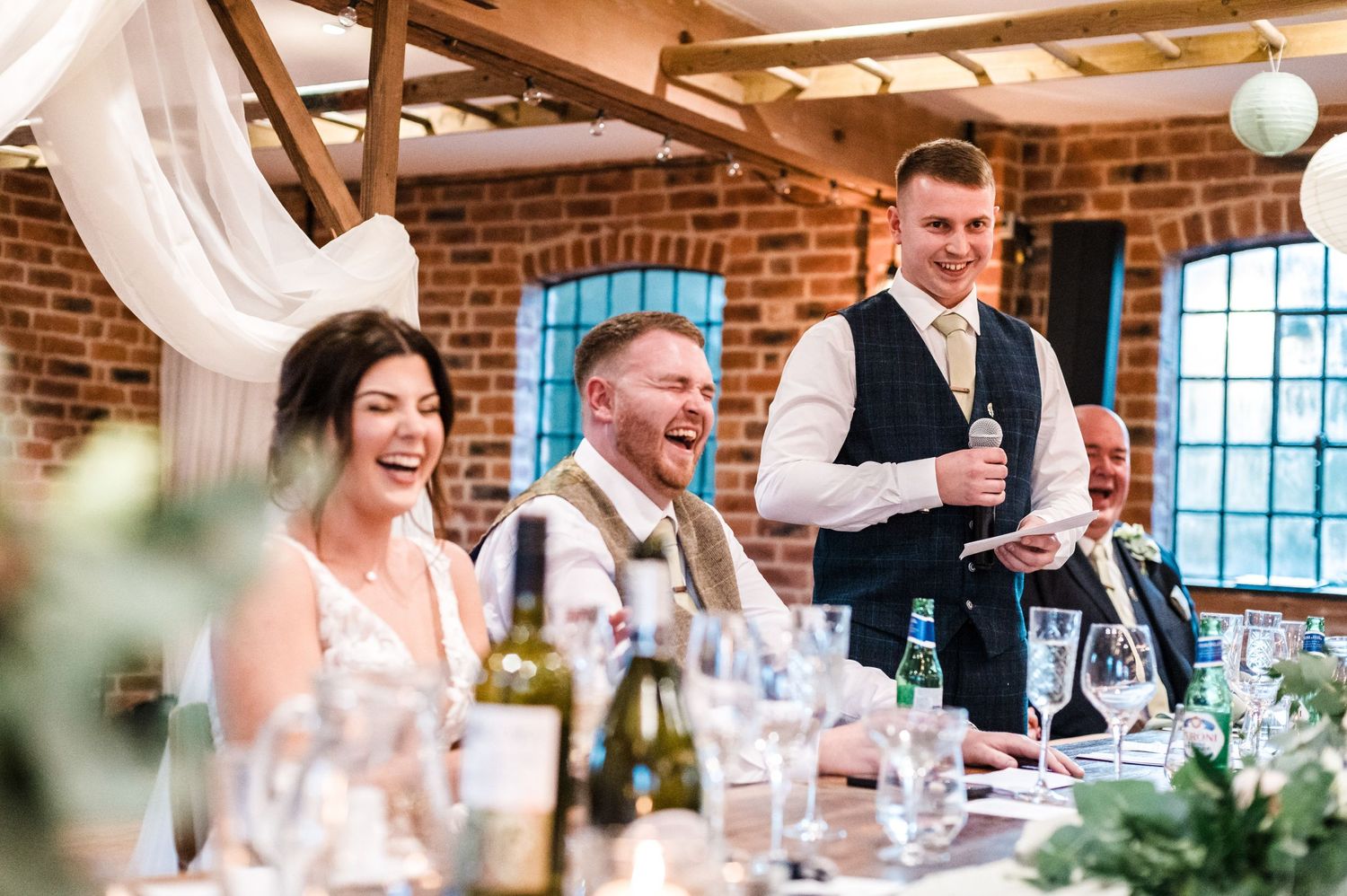 Joyful reactions during wedding speeches at rustic reception venue with exposed brick walls.