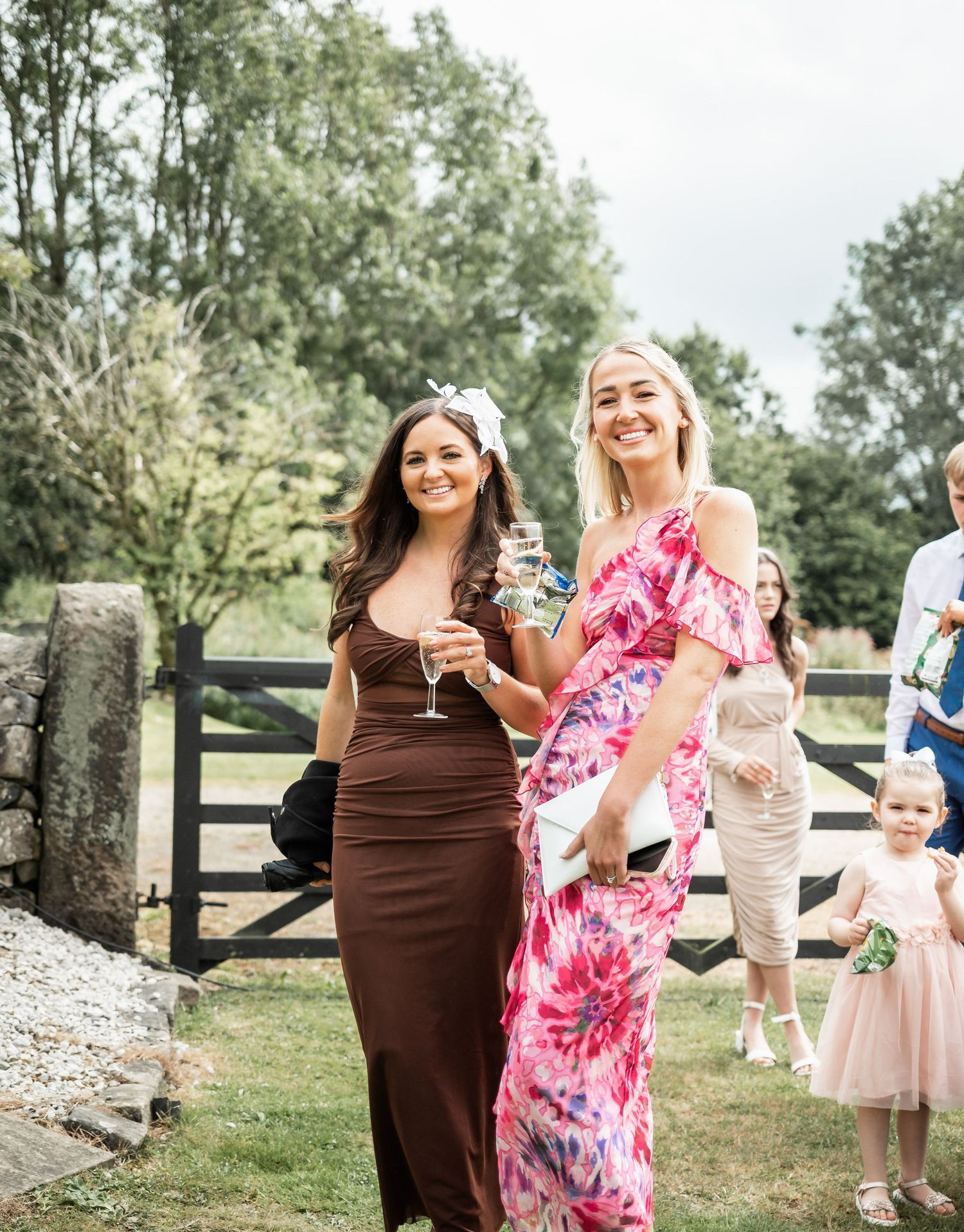 Two wedding guests in formal dresses, one brown and one pink floral, share a cheerful moment outdoors.