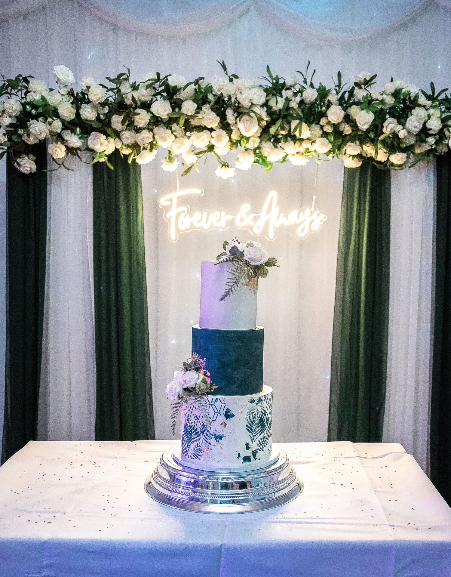 Elegant three-tier wedding cake with navy and white floral design under a lighted name sign and floral arch.