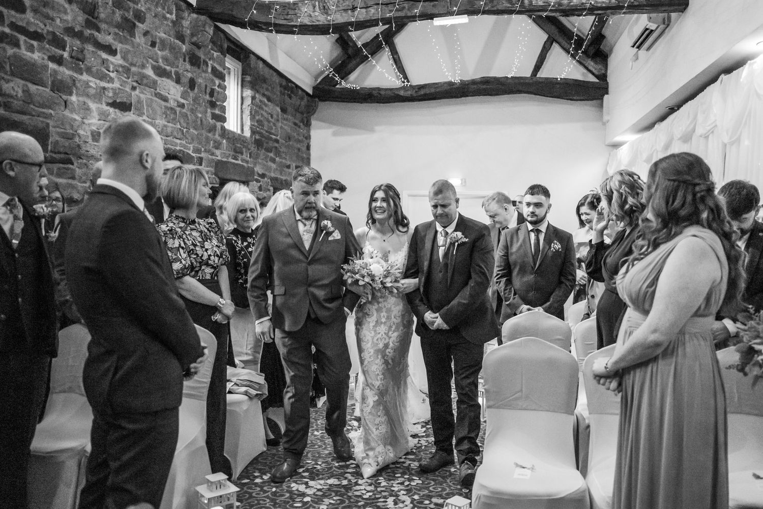 A wedding ceremony taking place in a rustic brick-walled venue with wooden beams and gathered guests in black and white.