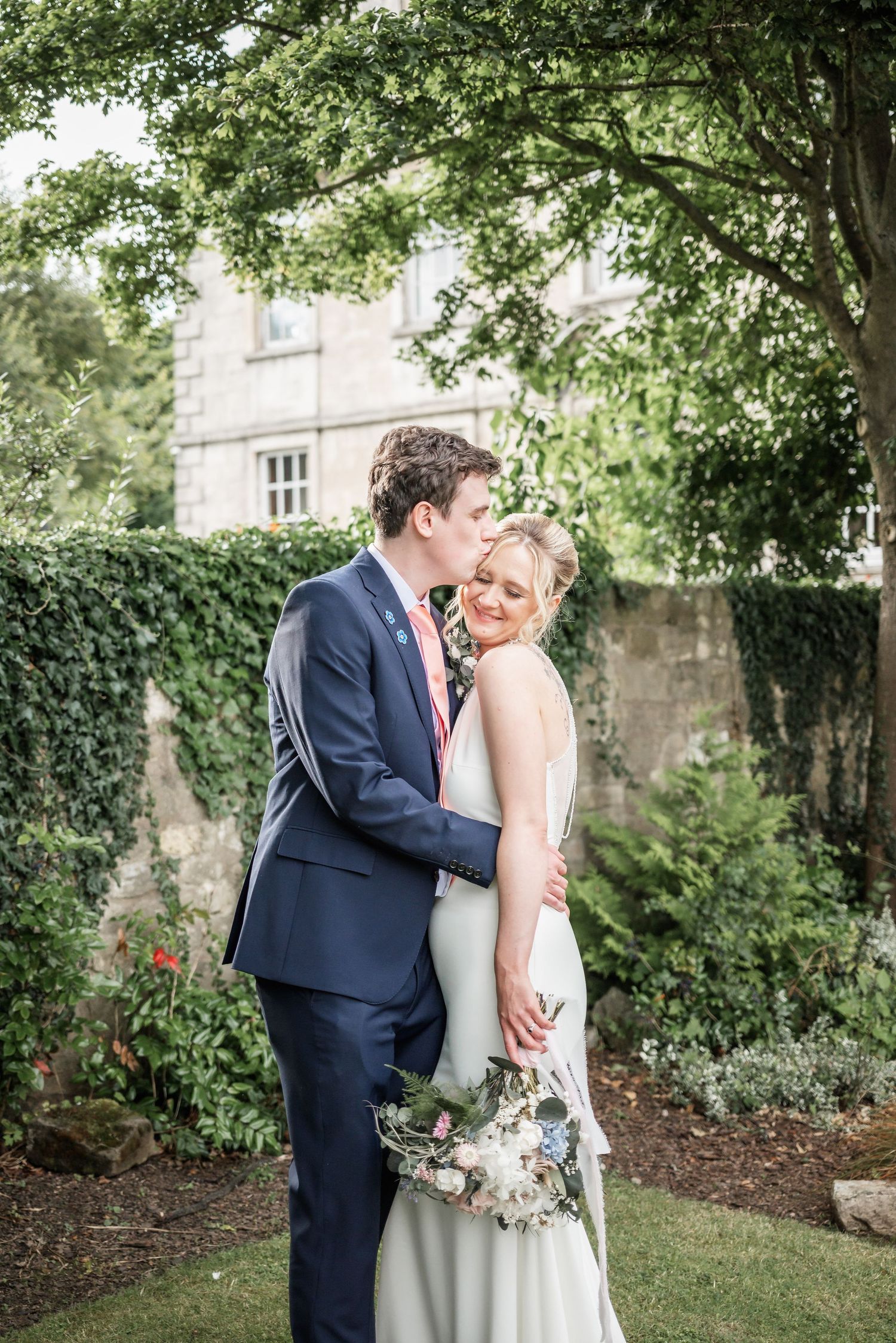 An intimate wedding portrait moment captured in a garden with historic building backdrop.