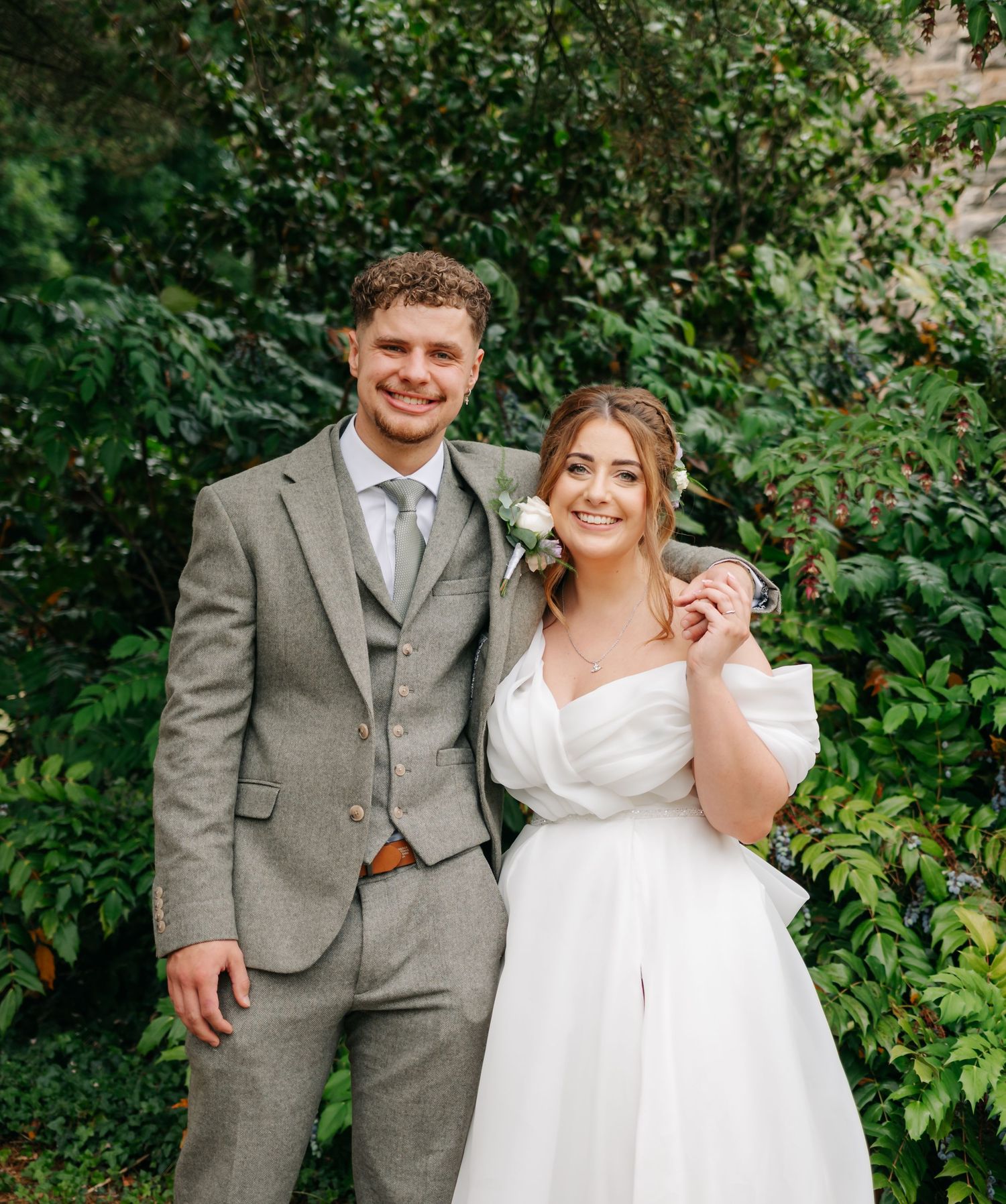 Happy newlyweds pose together in formal attire against a leafy green garden backdrop.